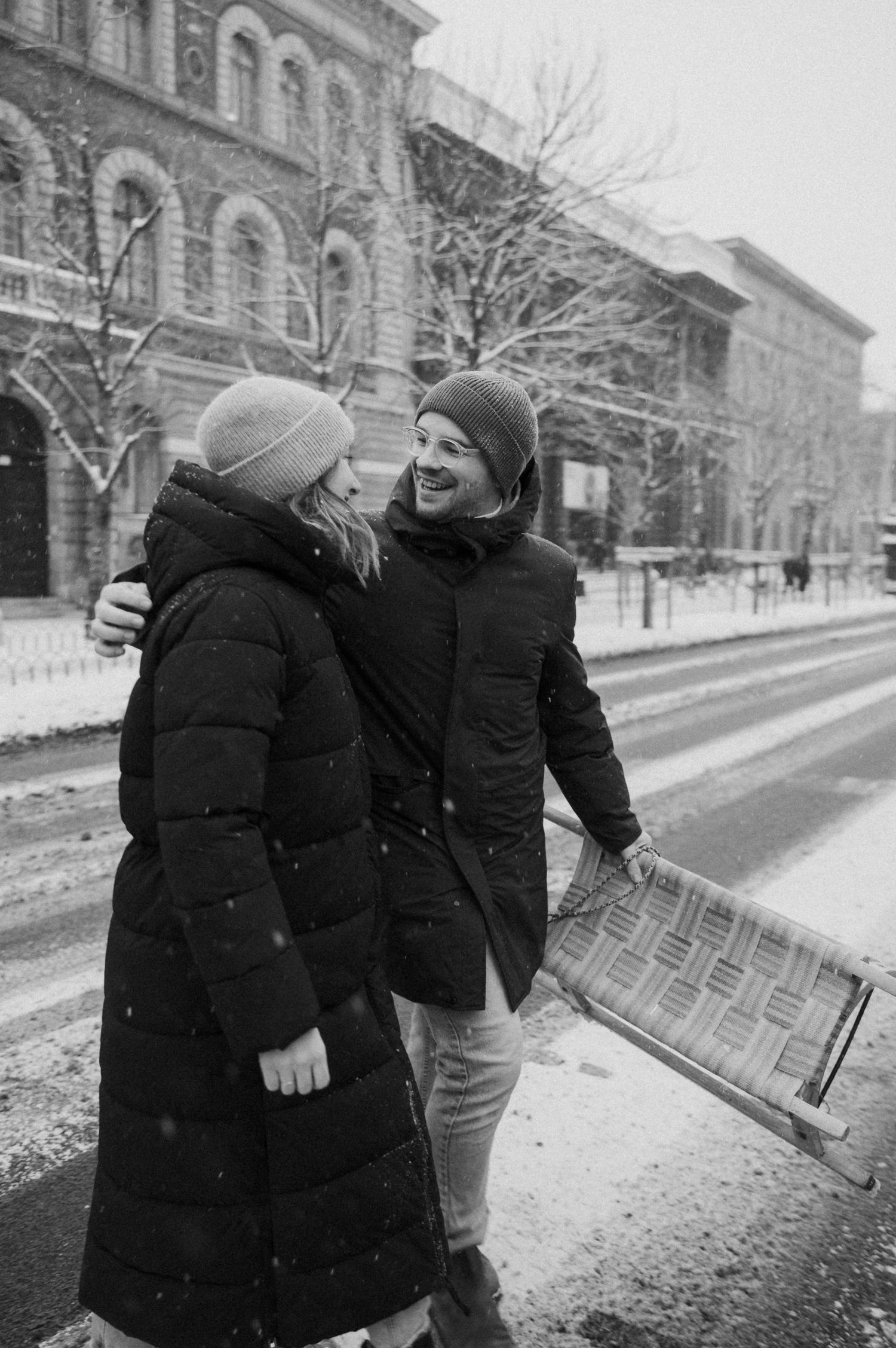 Two figures walking along a snowy street beside historic buildings.
