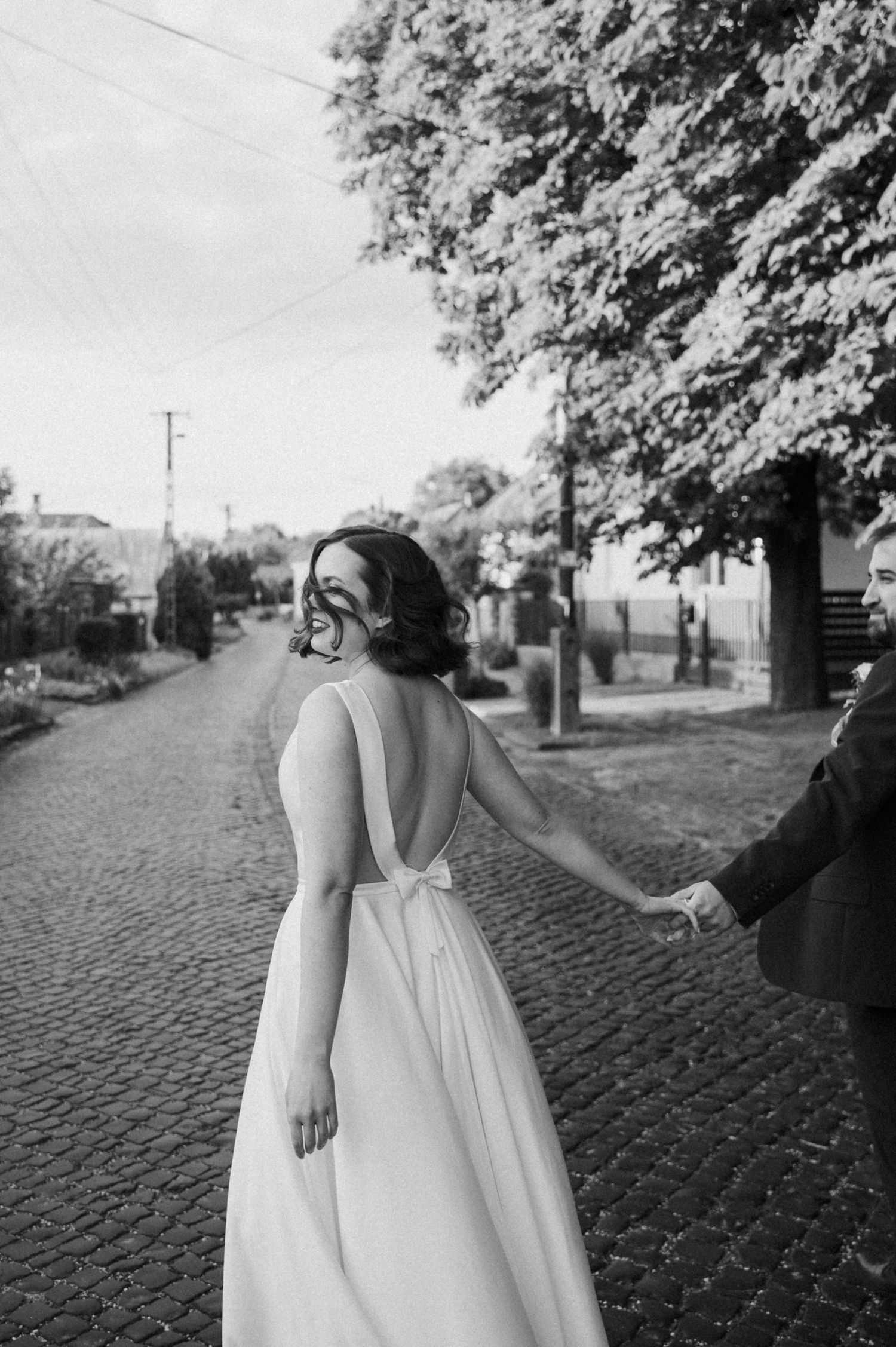 A person in a flowing white dress stands on a cobblestone path beneath leafy trees in a black and white photograph.