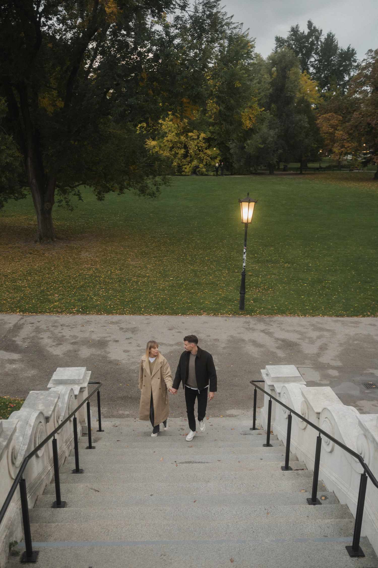A couple walking together on concrete steps with black railings in an autumn park setting with trees and lamp posts.
