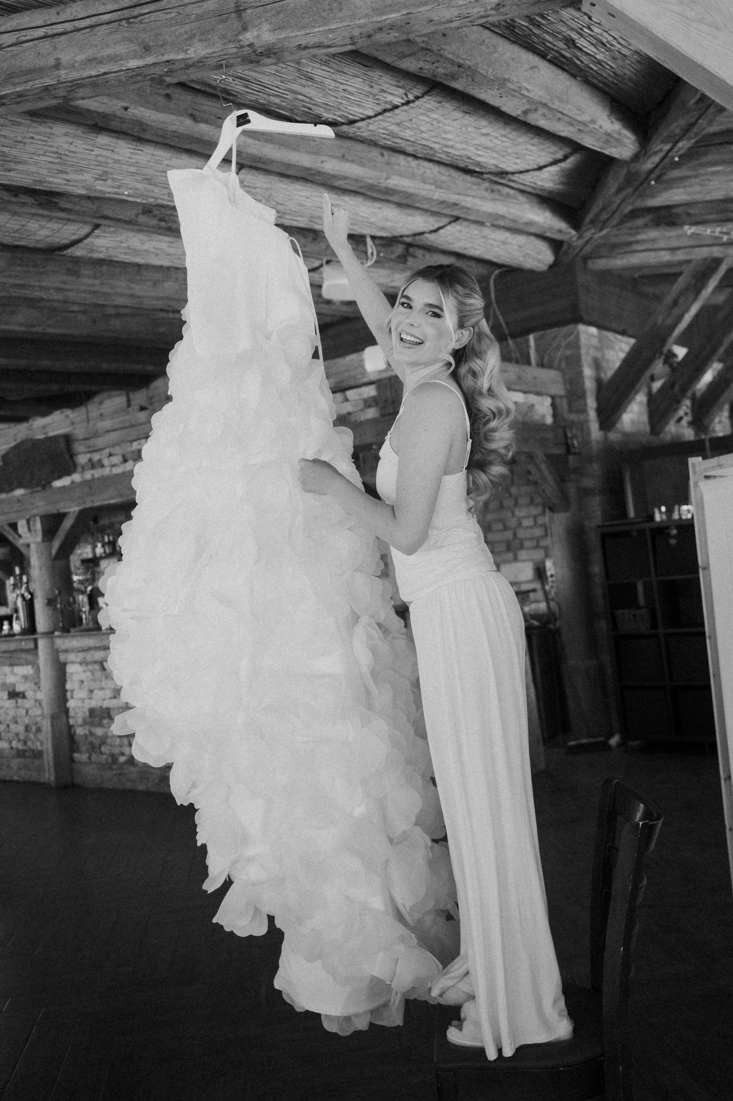 Bride in white gown joyfully holding her ruffled wedding dress in a rustic venue, black and white.