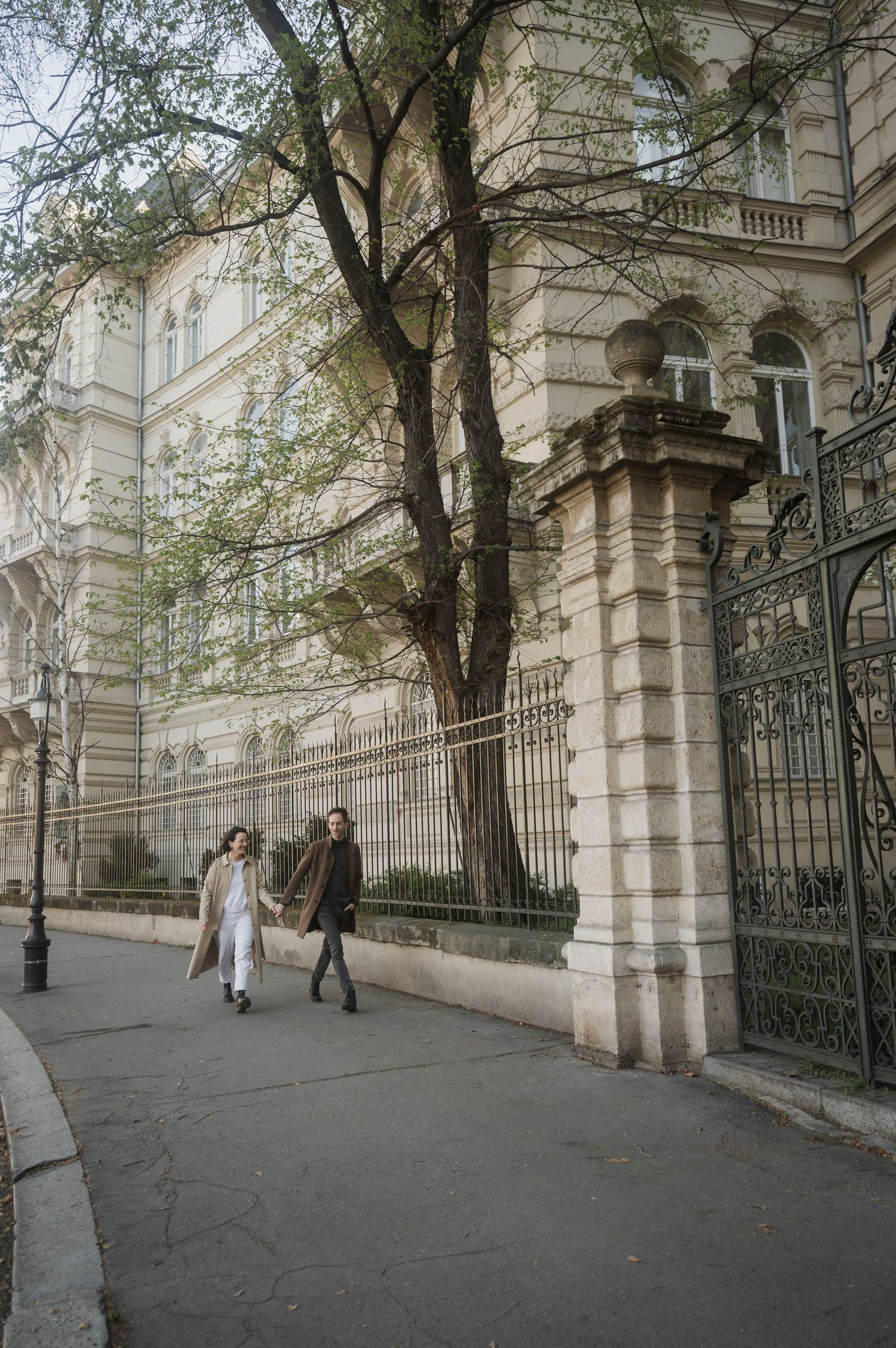 Two people walking along a sidewalk beside an ornate historic building with wrought iron gates and stone columns in autumn.