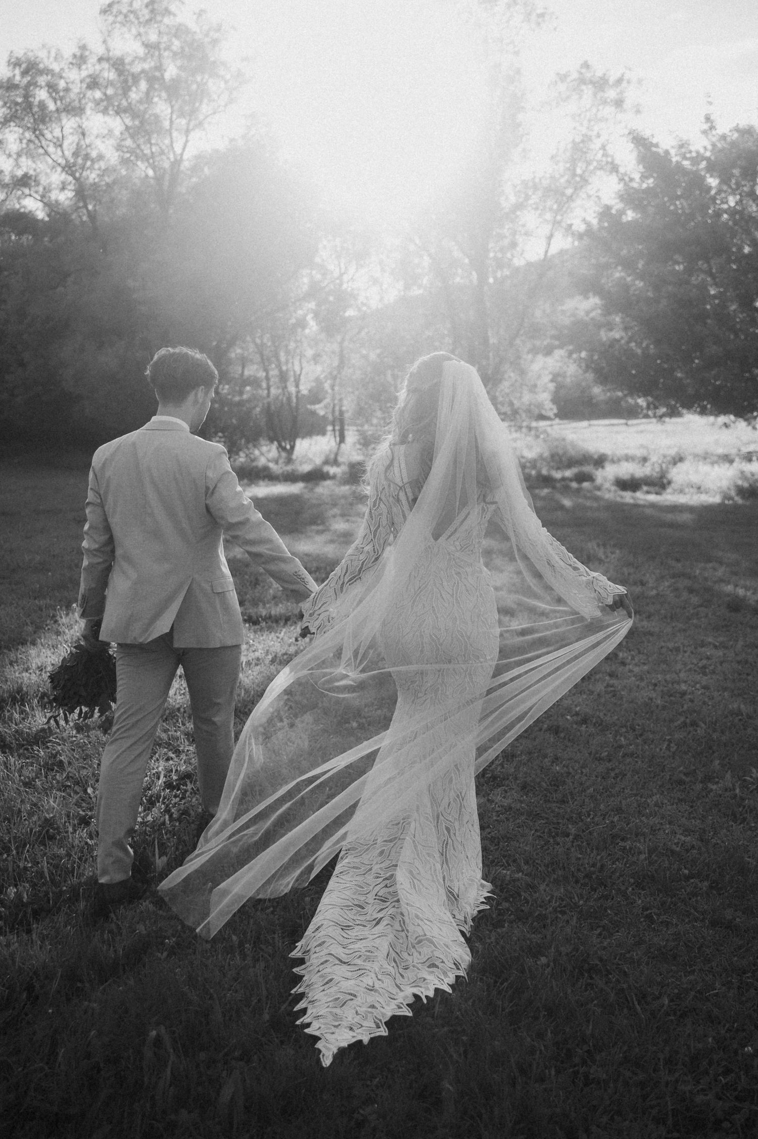 A romantic black and white wedding photo of a couple walking in sunlit field with flowing lace veil catching the breeze.