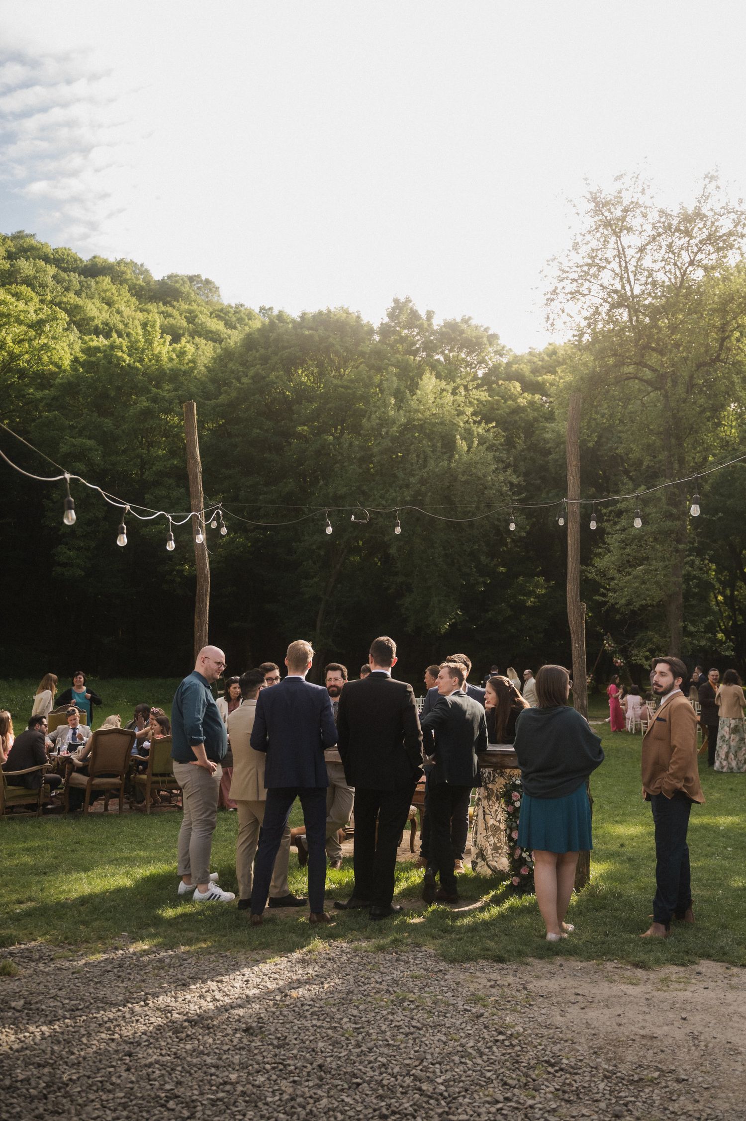 A group of guests gather outdoors at dusk under string lights during a social event in a wooded area.