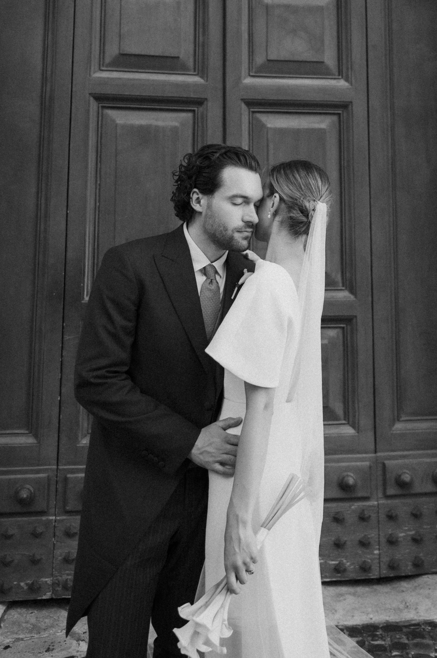Black and white wedding portrait showing tender moment against architectural backdrop.