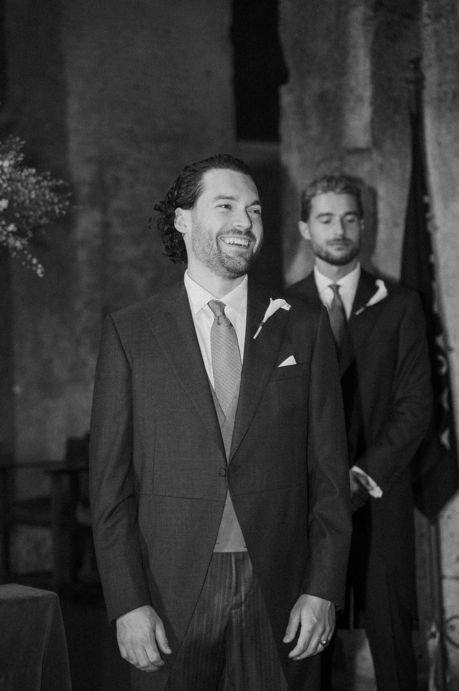 A groom in formal attire smiles during a wedding ceremony in a historic brick venue.