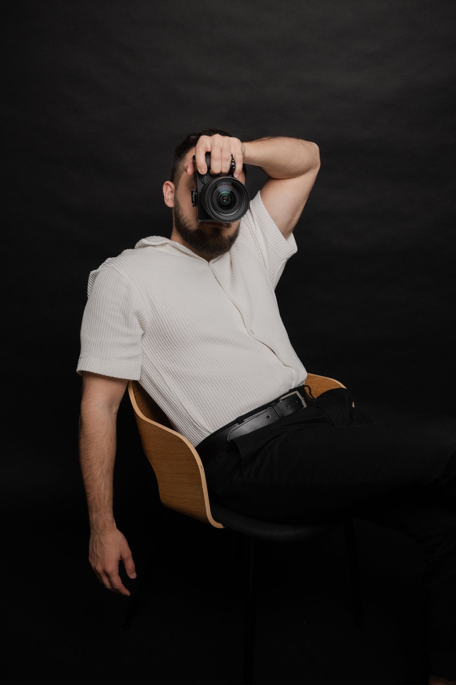 A photographer in a white polo shirt sits on a wooden chair against a black backdrop while taking a photo.
