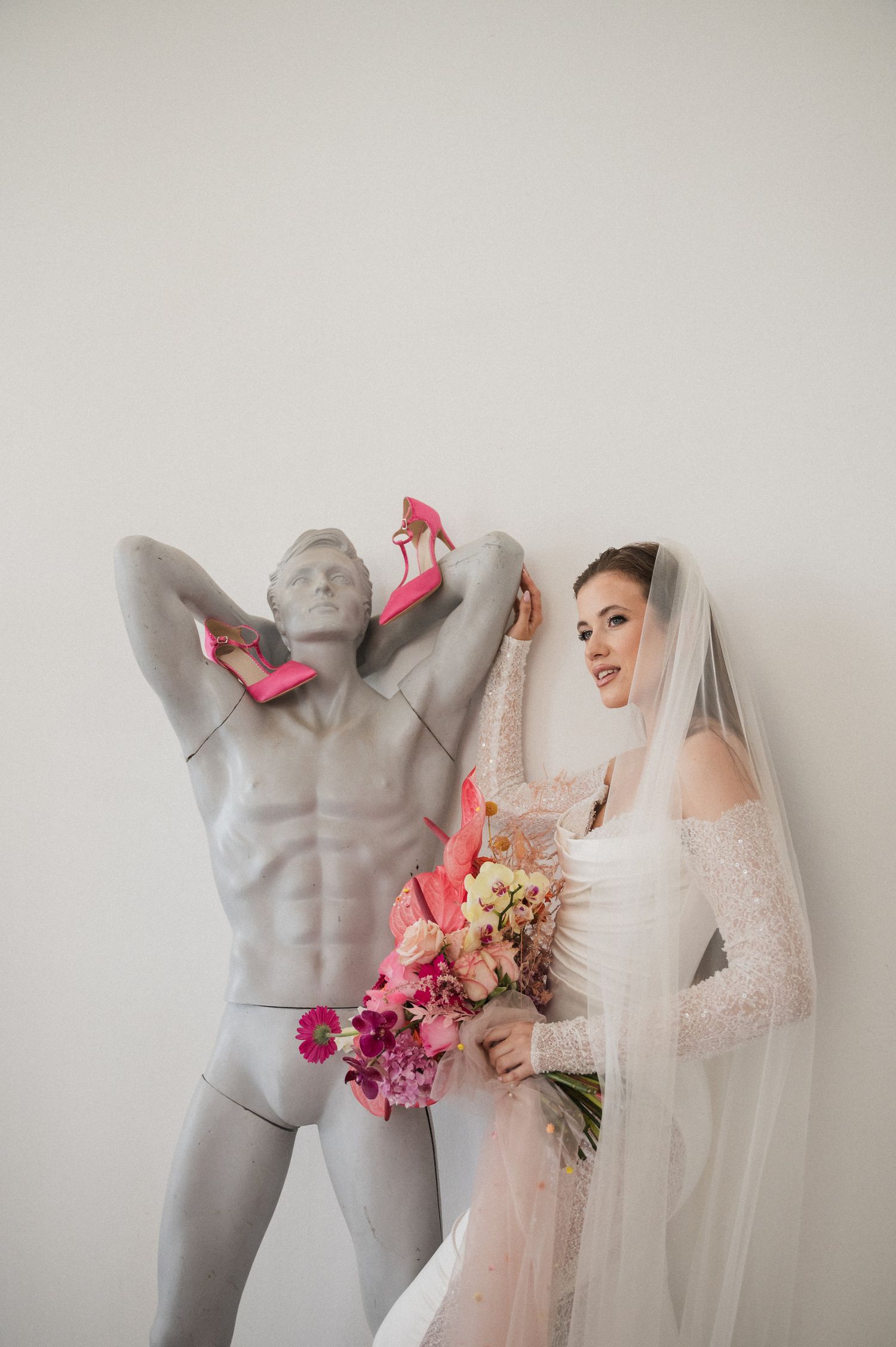Whimsical wedding photo with bride posing next to decorative statue adorned with pink shoes.