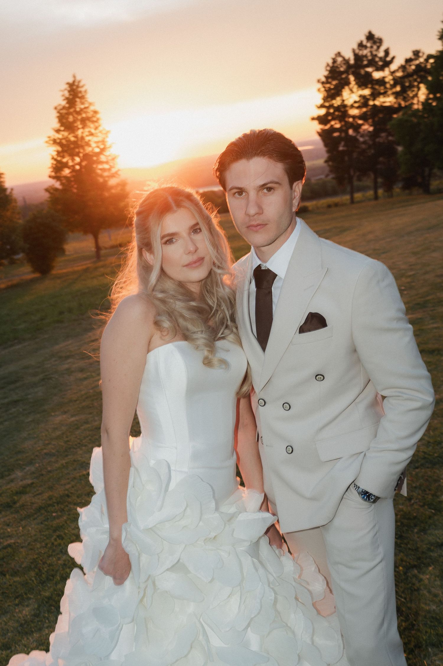 Elegant bride and groom posing together at sunset in white ruffled gown and cream double-breasted suit.