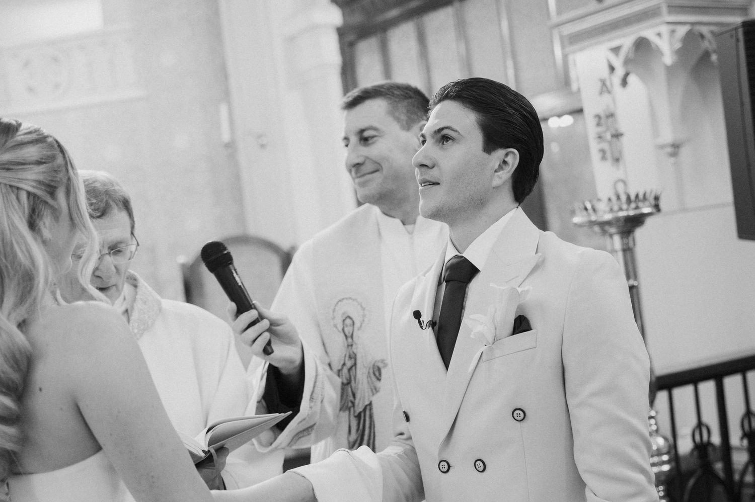 Groom in white suit smiling during wedding ceremony vows inside a church, black and white photo.