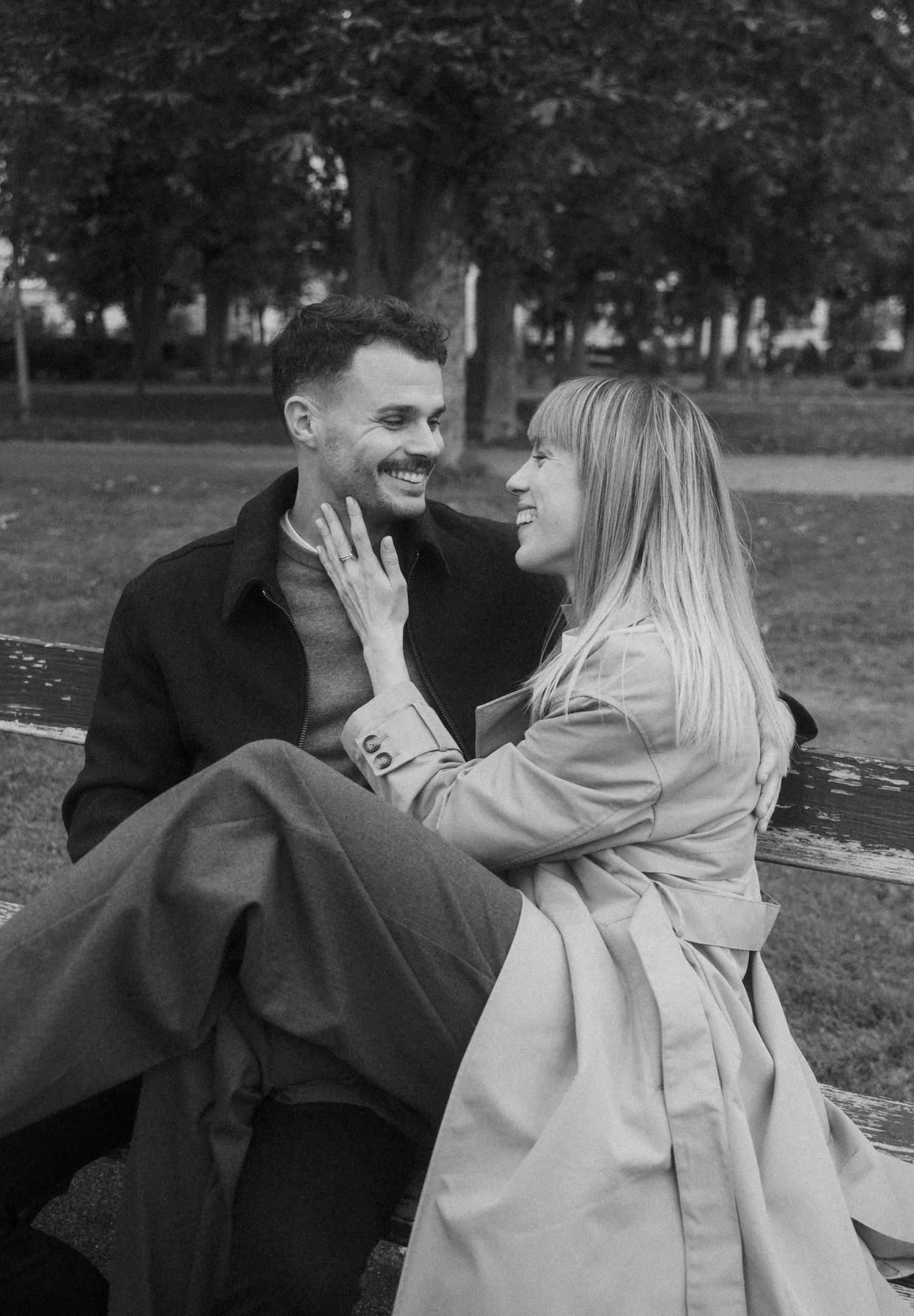 A romantic black and white photo of a couple laughing and embracing on a bench in a park.