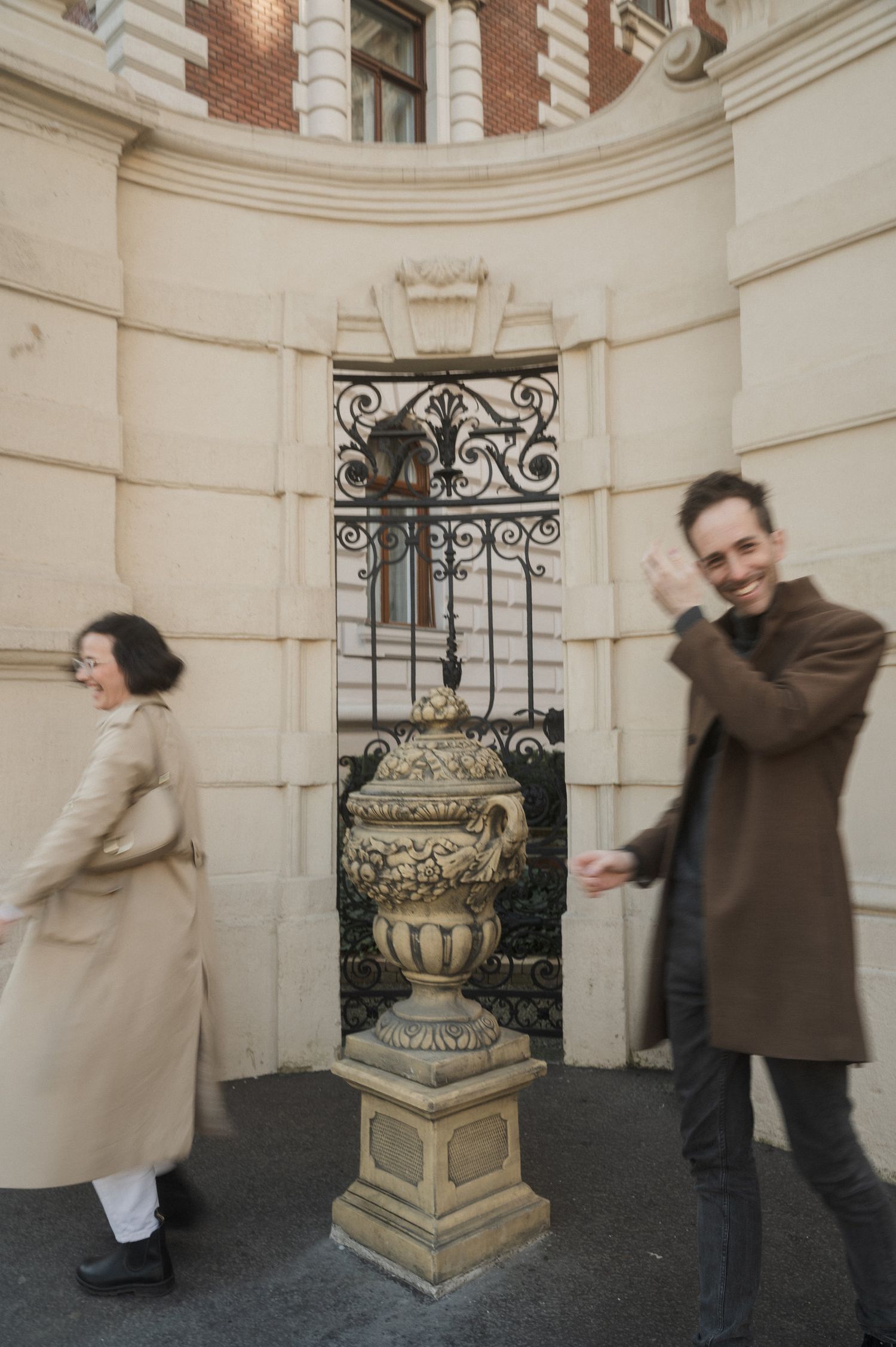 Two people playfully pose in front of a curved stone wall with decorative iron grates and a stone planter.