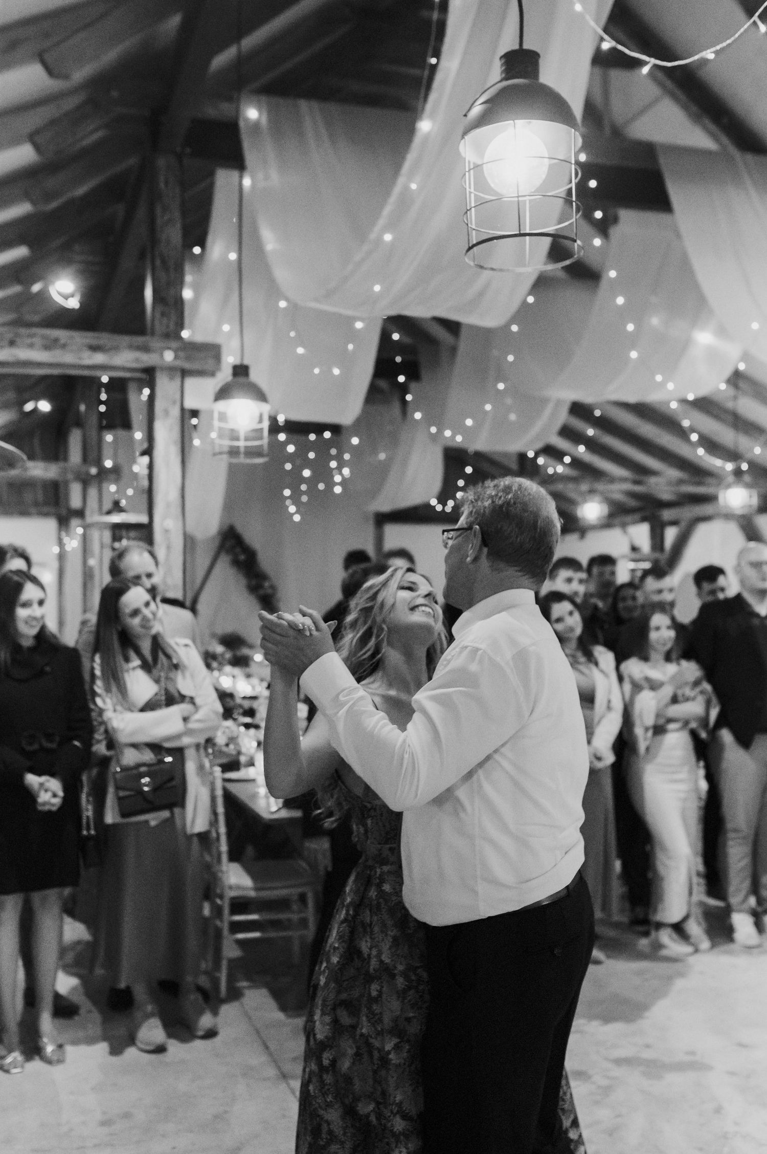 People dancing at a wedding reception under twinkling string lights in a rustic indoor venue.