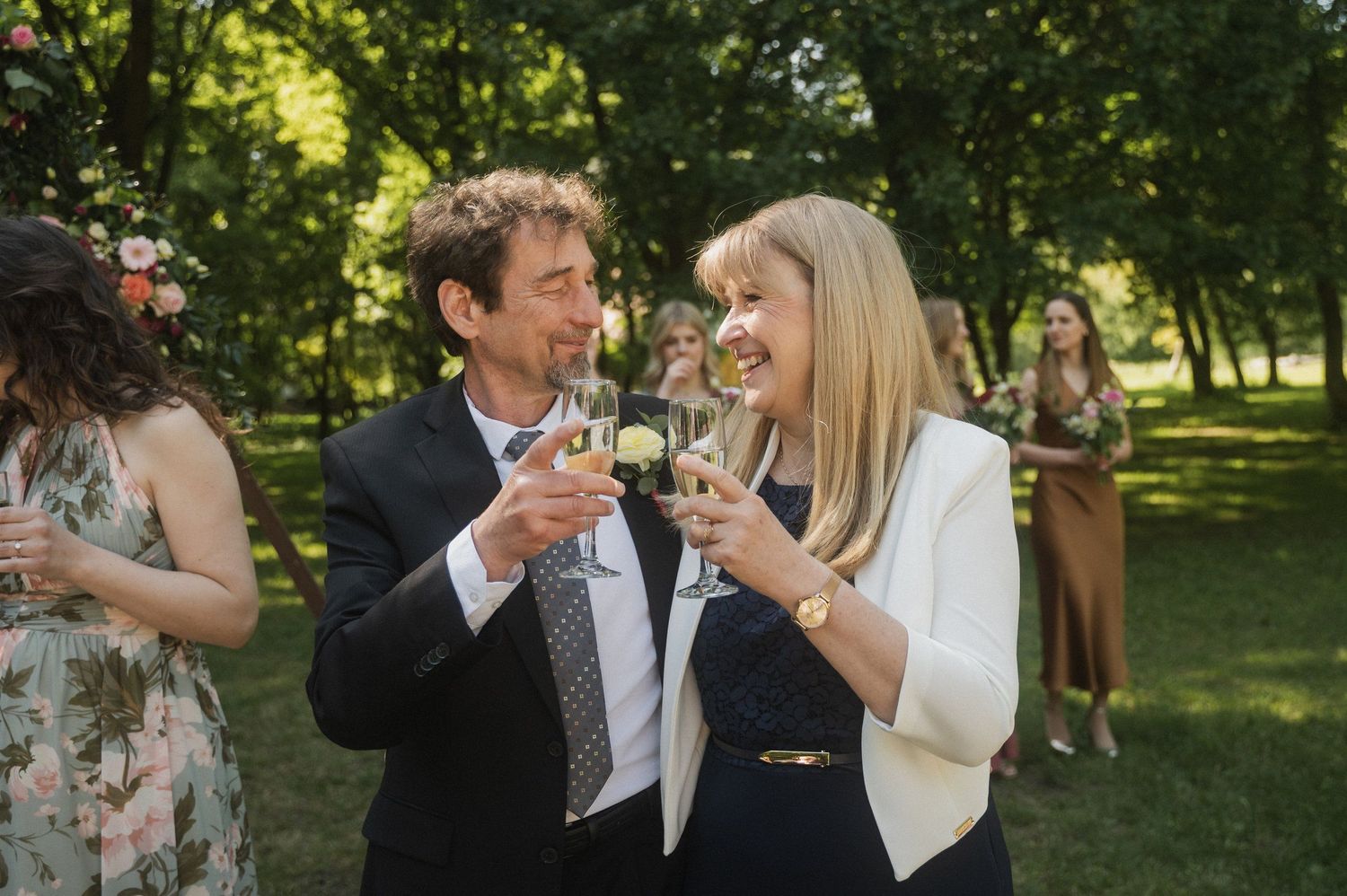 A couple toasts with champagne glasses during an outdoor wedding celebration in a sunlit garden setting.