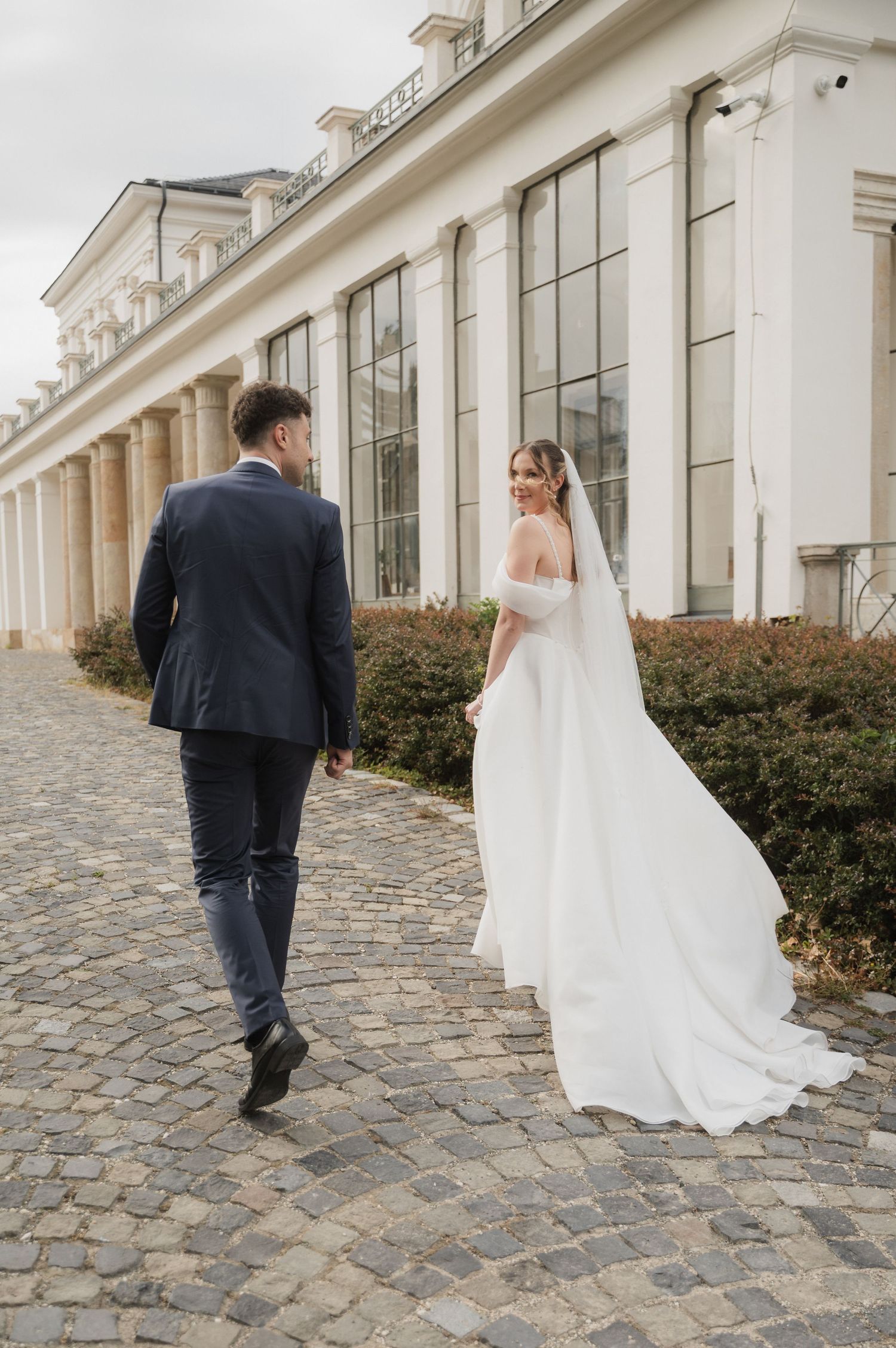 Bride and groom walking along cobblestone path beside elegant white building on their wedding day.