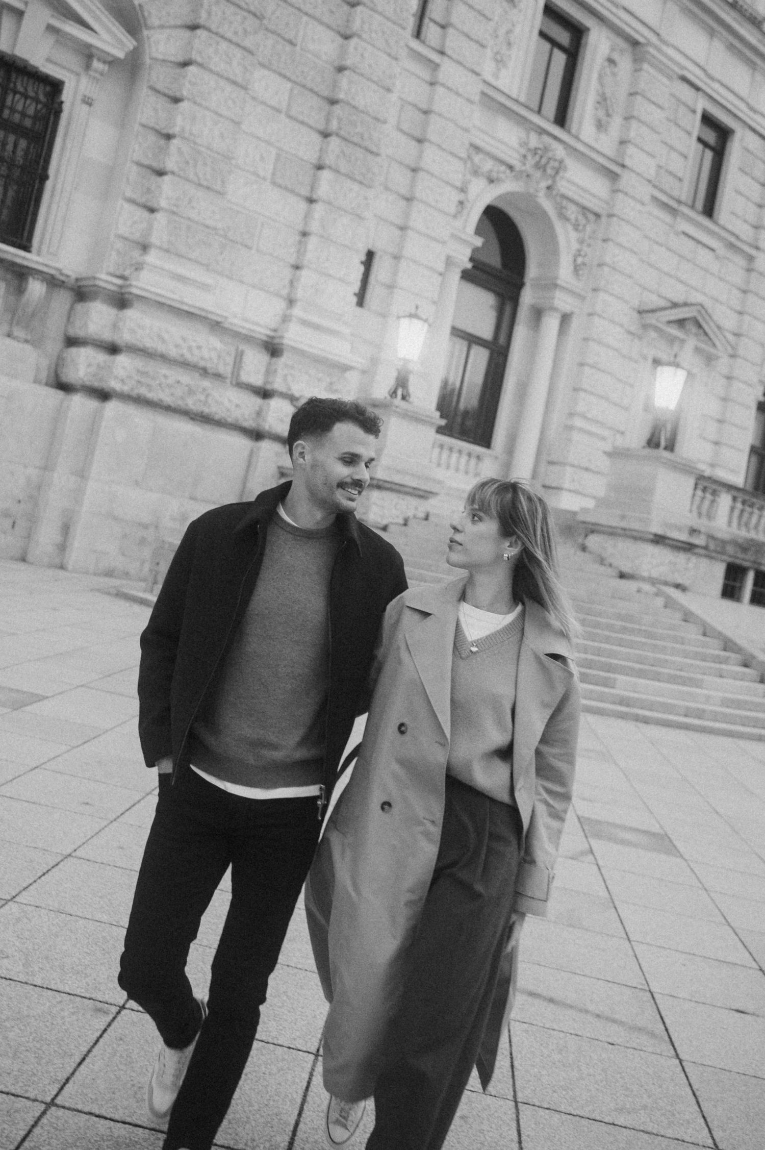 A couple in casual winter attire sharing a romantic moment outside a grand stone building with arched architecture.