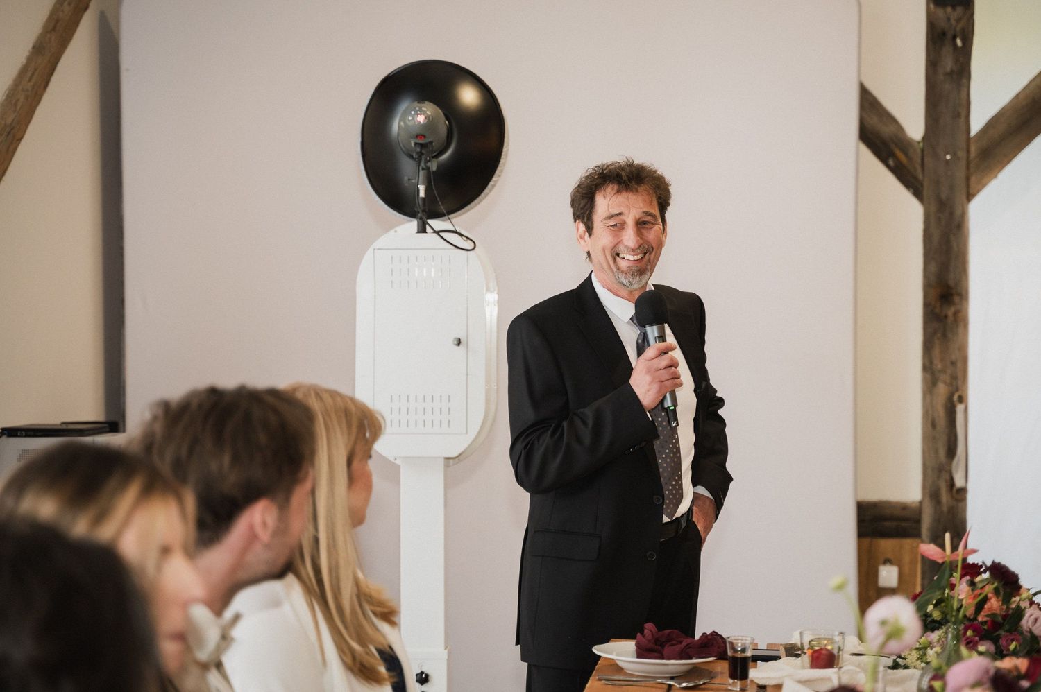 Wedding reception guests laugh and smile during emotional speeches at a rustic barn venue.