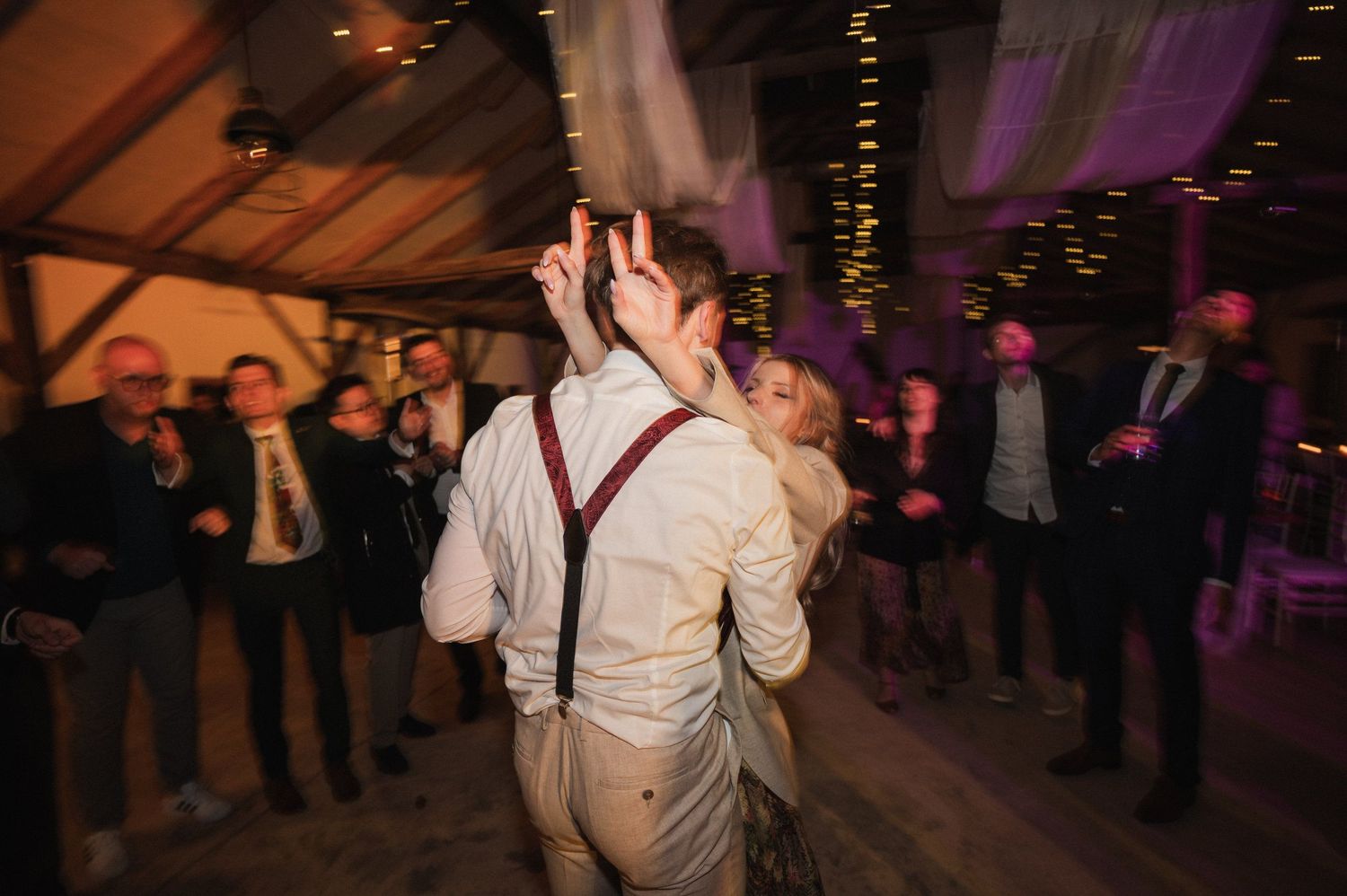 A couple dances together at a wedding reception with purple lighting and guests watching in the background.