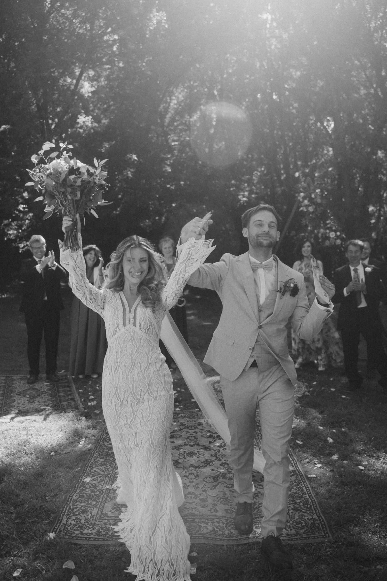 A newlywed couple celebrates joyfully in an outdoor garden setting with natural sunlight filtering through trees.