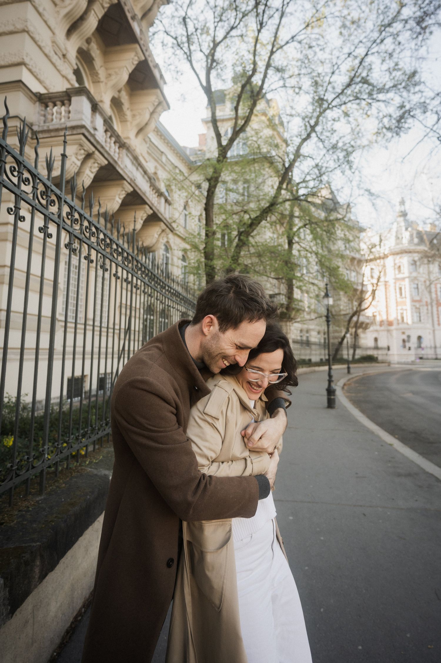 A couple embraces on a romantic street in Paris with elegant architecture and iron fencing in the background.