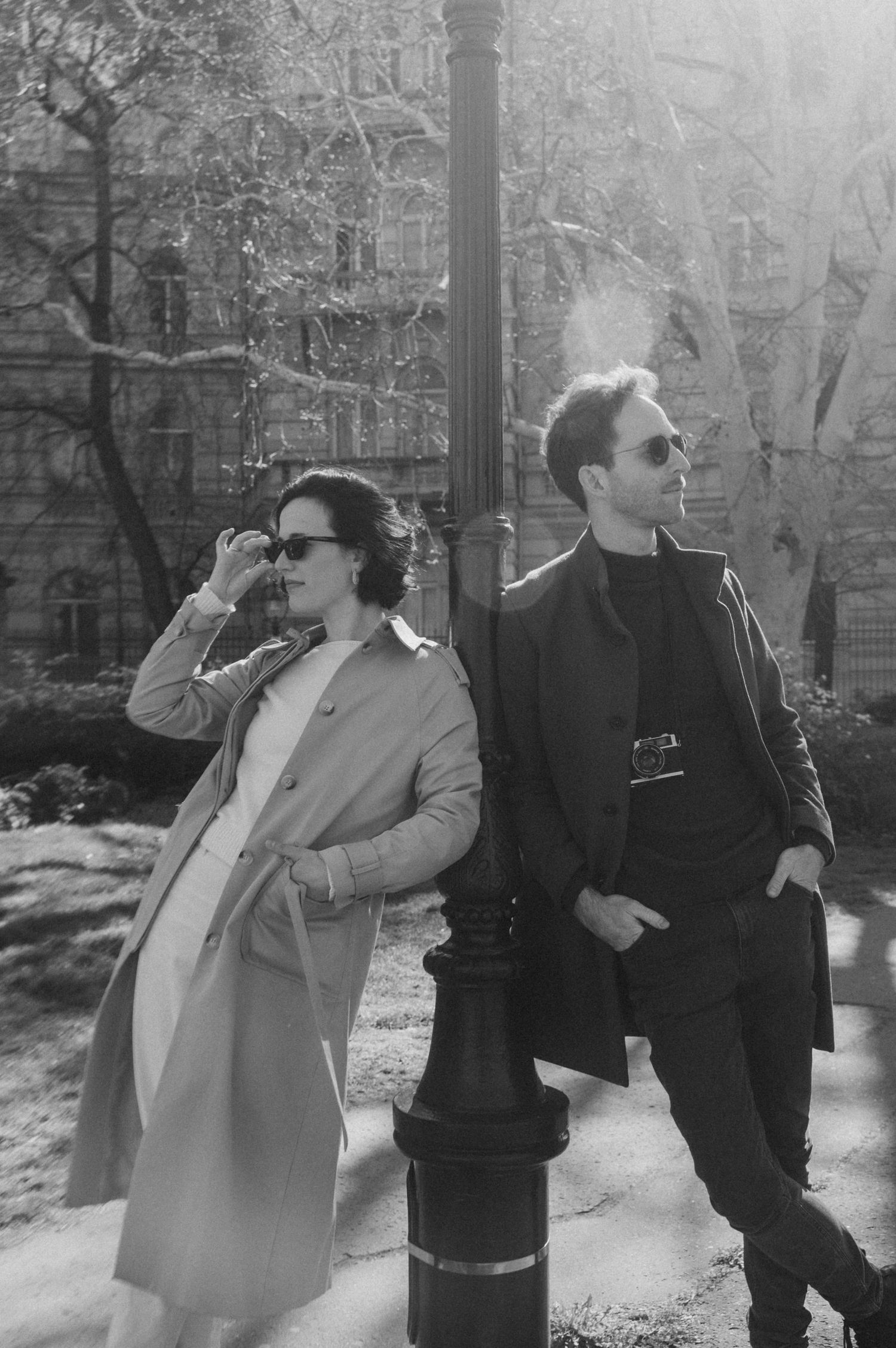 Two people in sunglasses and coats stand by a lamppost on a misty day in what appears to be a park setting.