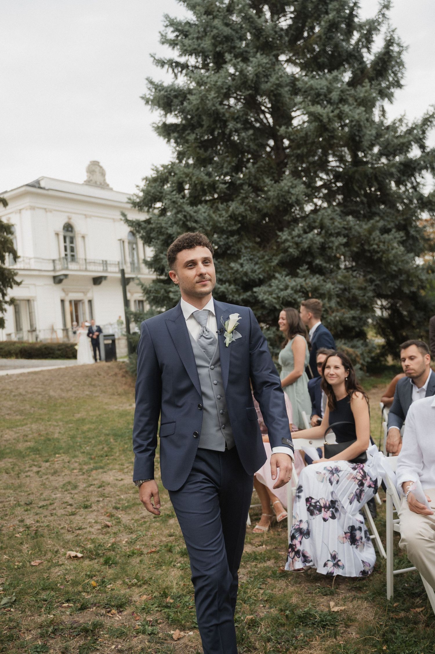 Groom in navy blue suit walking down outdoor wedding aisle with guests seated beside him.