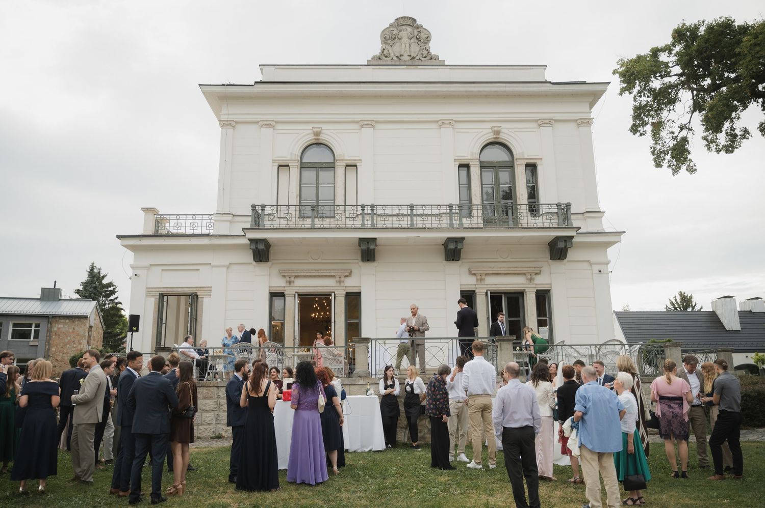 Wedding guests mingle on lawn outside elegant white neoclassical villa during outdoor reception.