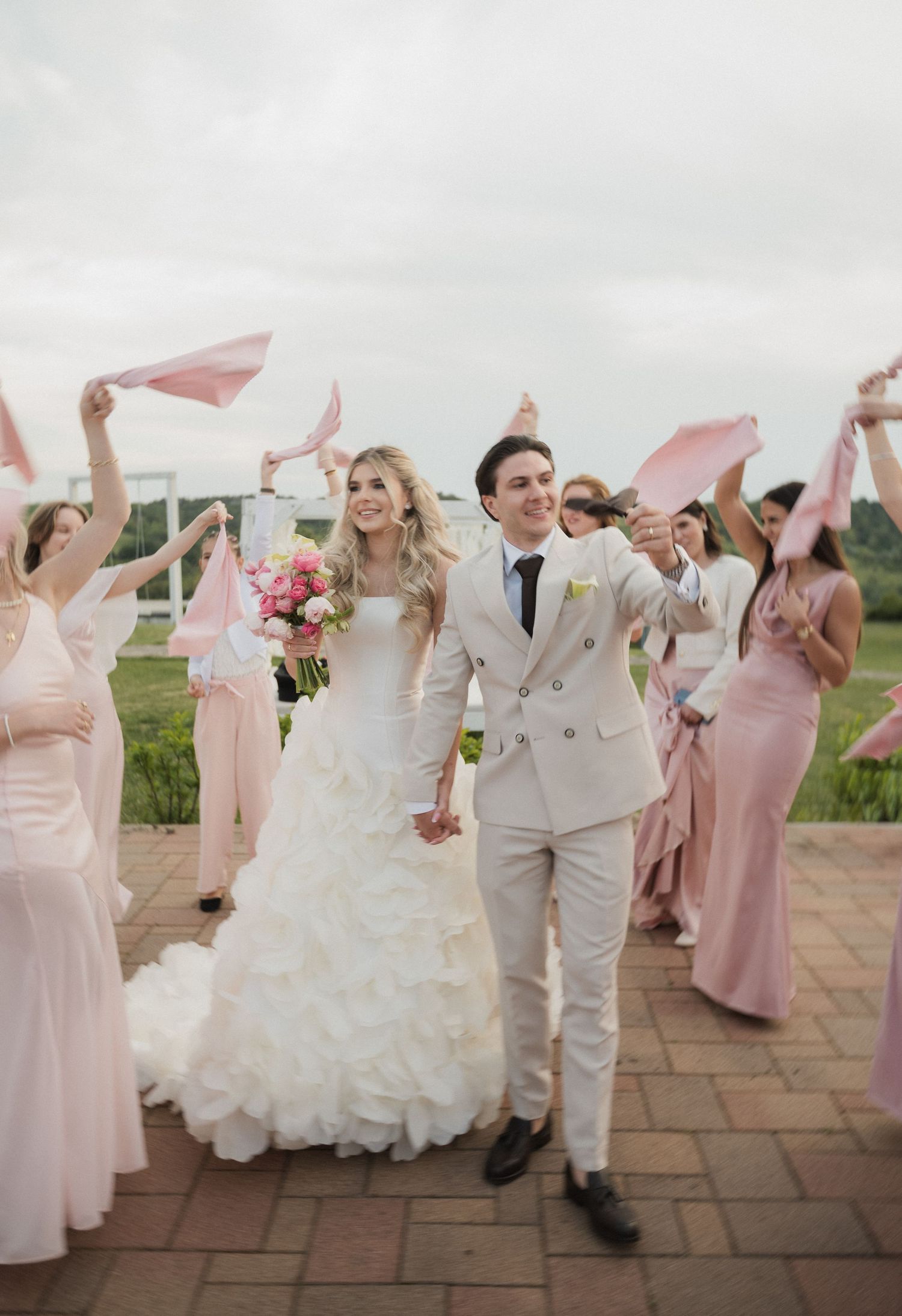 Bride and groom walk joyfully as bridesmaids in pink wave ribbons at outdoor wedding.