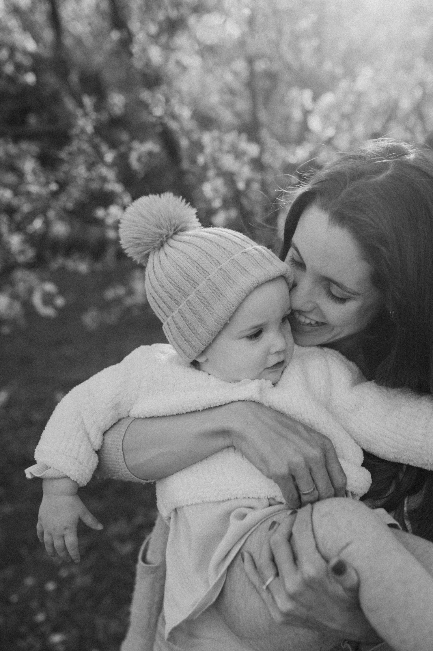 Black and white photo of a smiling mother kissing her baby wearing a knit pom-pom hat outdoors.