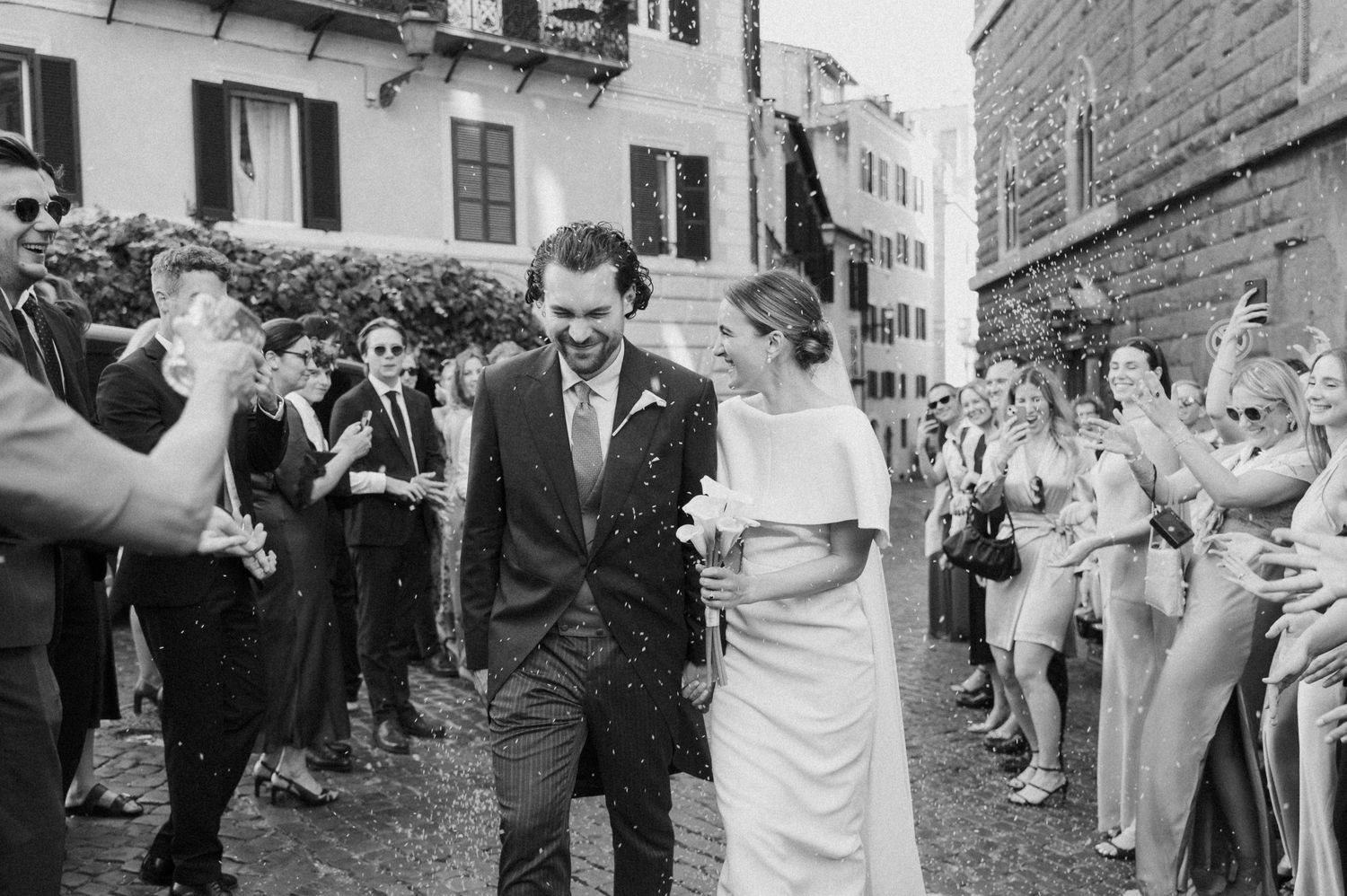 Joyful wedding procession with guests celebrating on European cobblestone street.