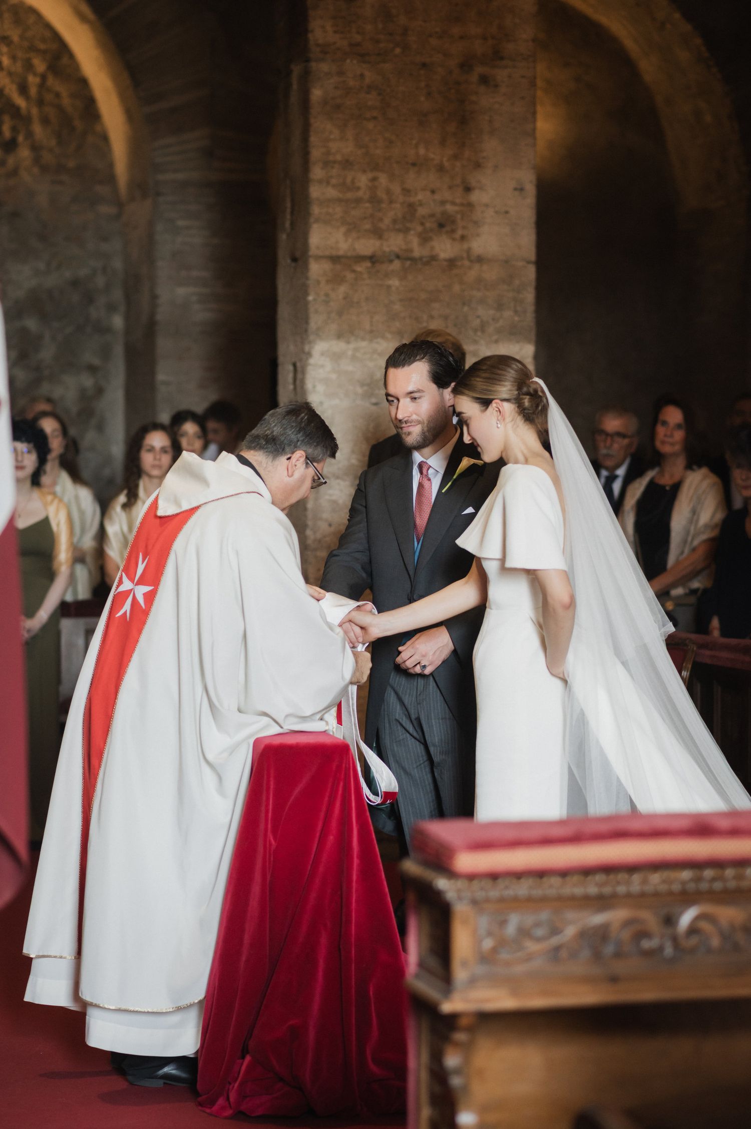 Religious wedding ceremony with clergy in white robes and red accents in a historic stone church.