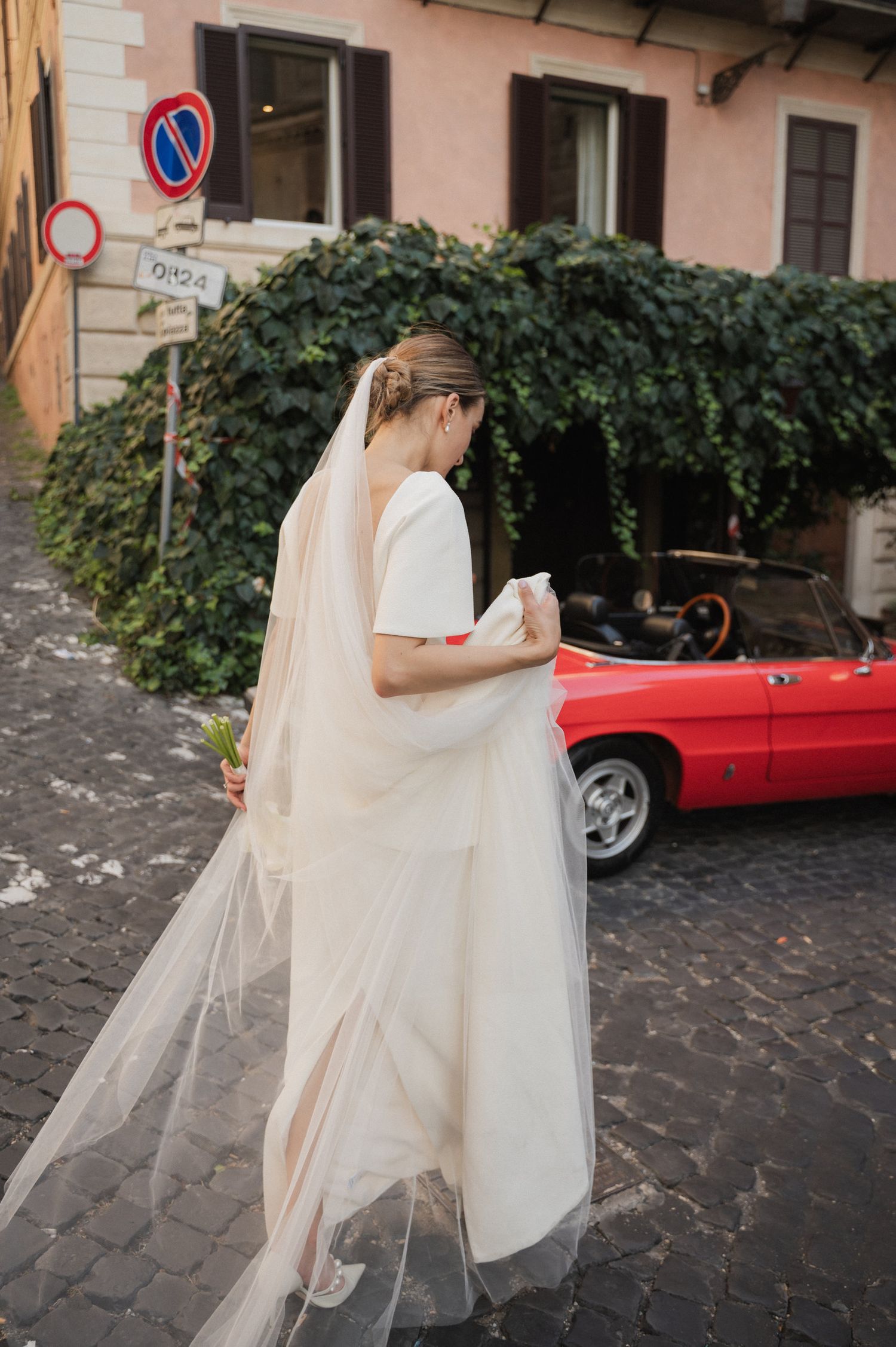 Bride in flowing white dress walks past vintage red car on cobblestone street.