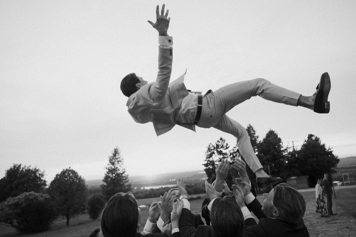 Black and white photo of groom being tossed in the air by wedding guests outdoors.