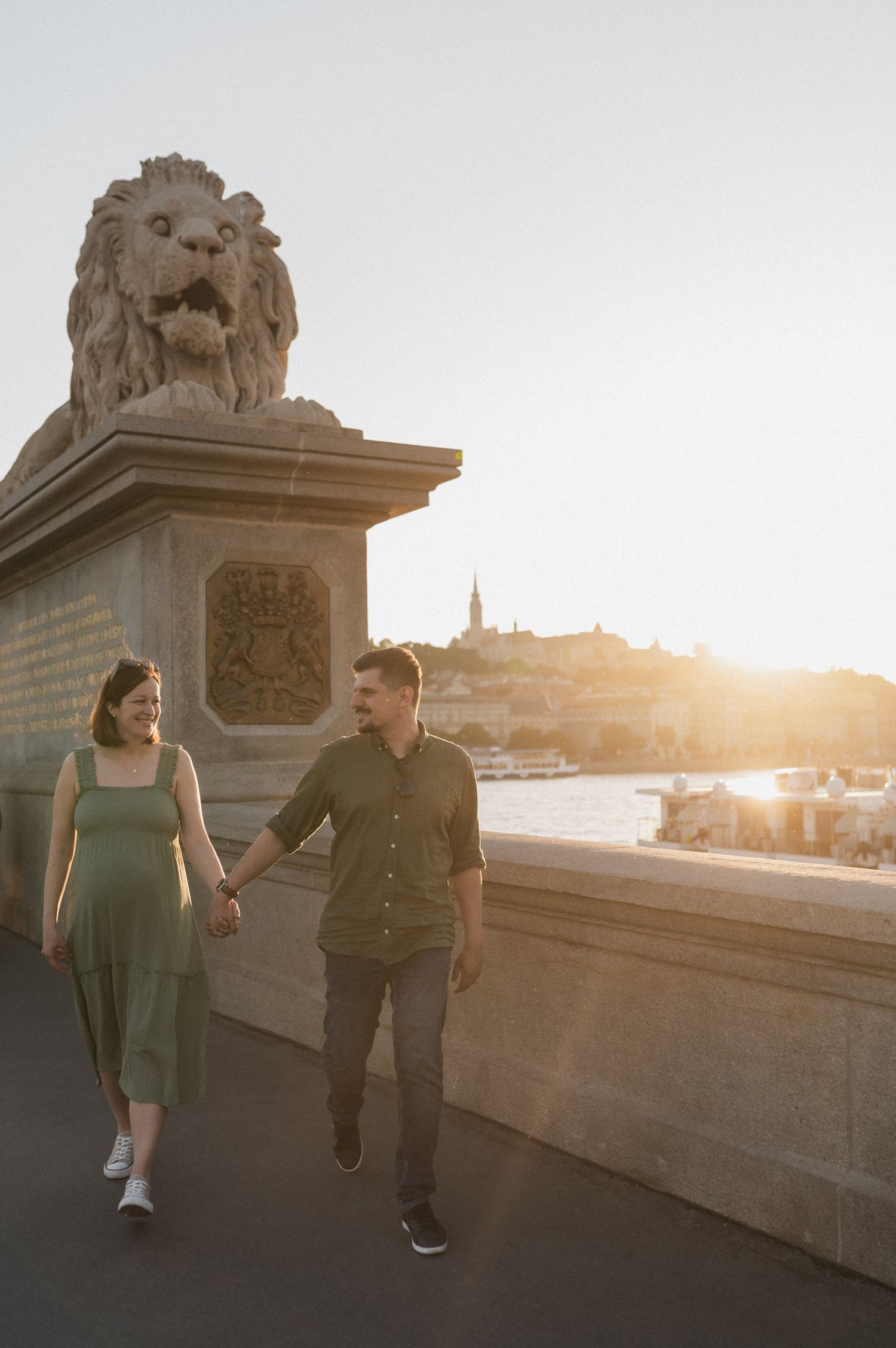 Couple walking hand in hand at sunset on Chain Bridge in Budapest with iconic lion statue and city skyline in background.