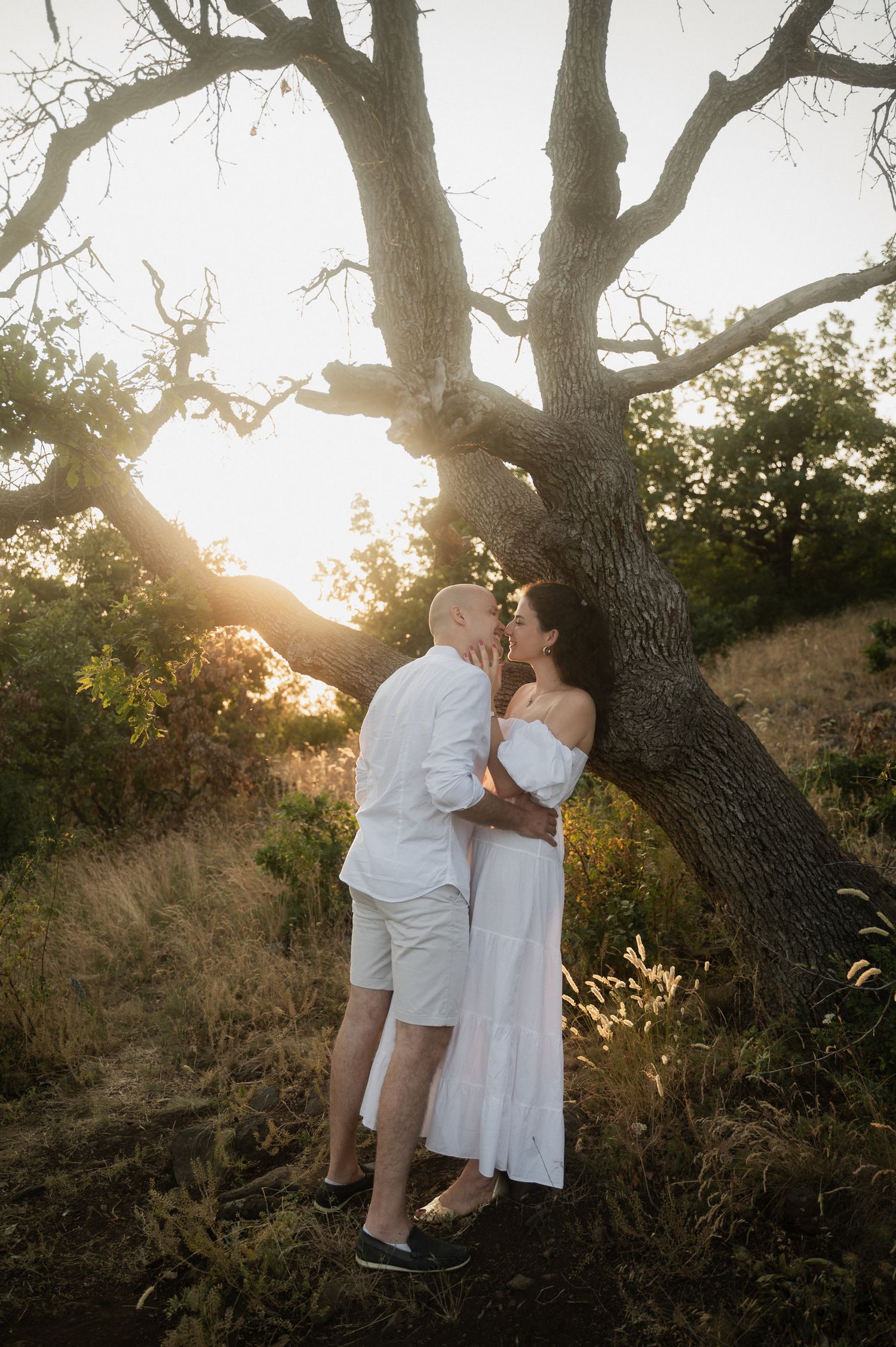 A couple shares a romantic moment at sunset under a large oak tree in a natural outdoor setting.