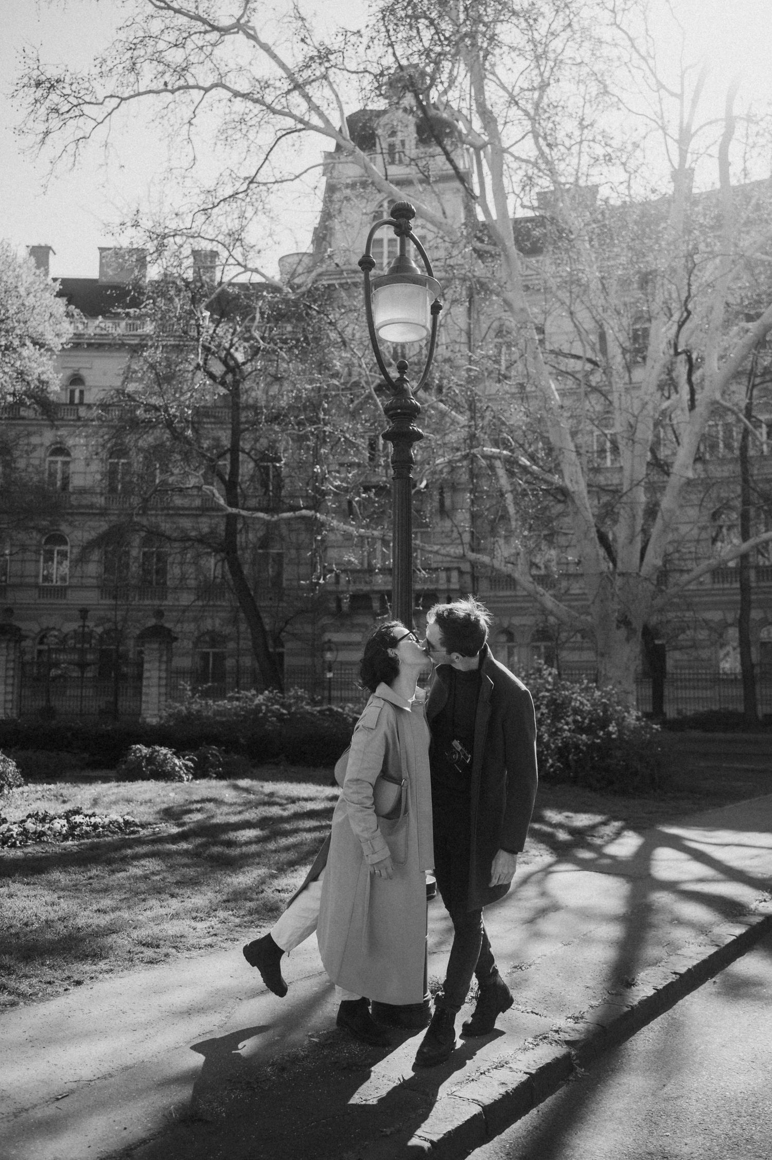 A couple walking together through a park on a sunny day casting long shadows on the ground.