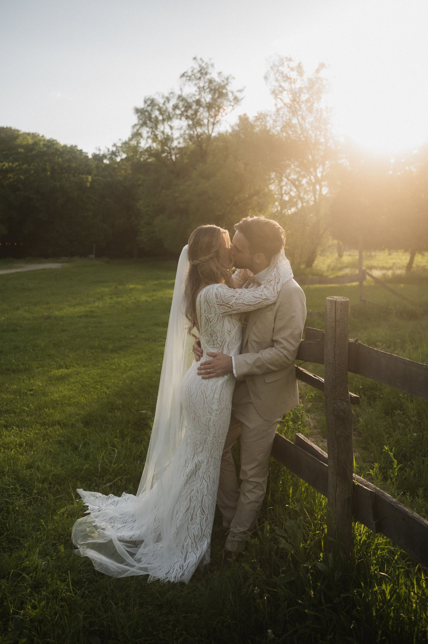 Romantic wedding couple embraces in golden sunlit field at sunset with flowing white dress and veil.
