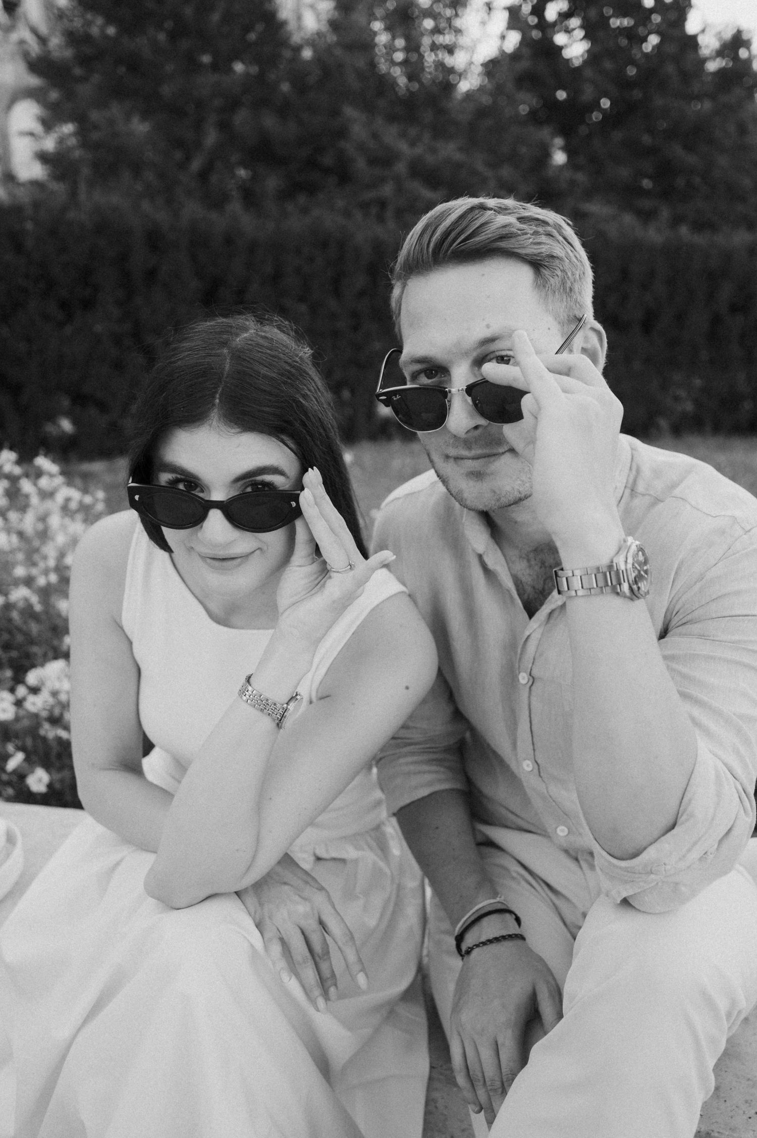 Black and white photo of a stylish couple sitting outdoors, both wearing sunglasses and white outfits, posing confidently.