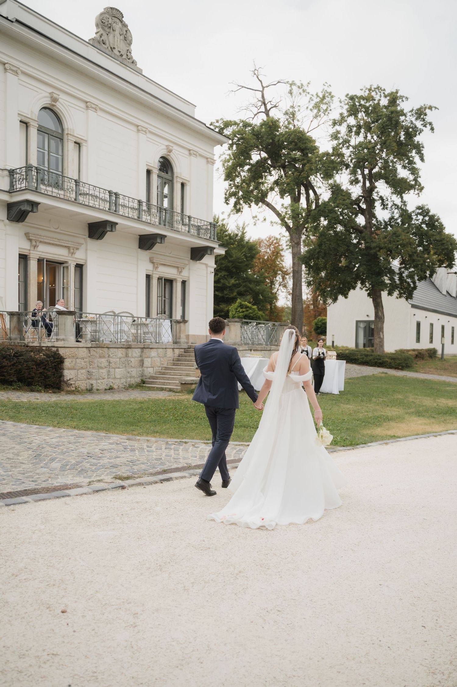 Bride in cathedral veil and groom in navy suit walk hand in hand toward elegant white wedding venue.