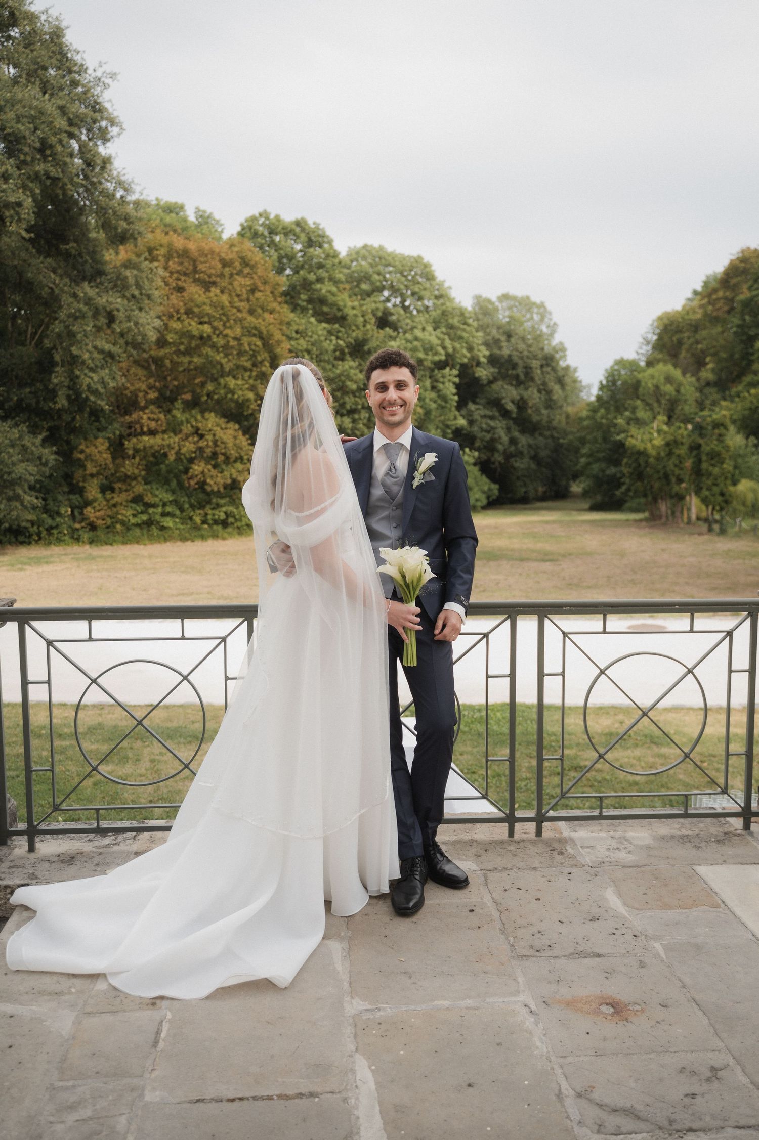 Smiling groom holds bouquet beside veiled bride on stone terrace with lush green park backdrop.