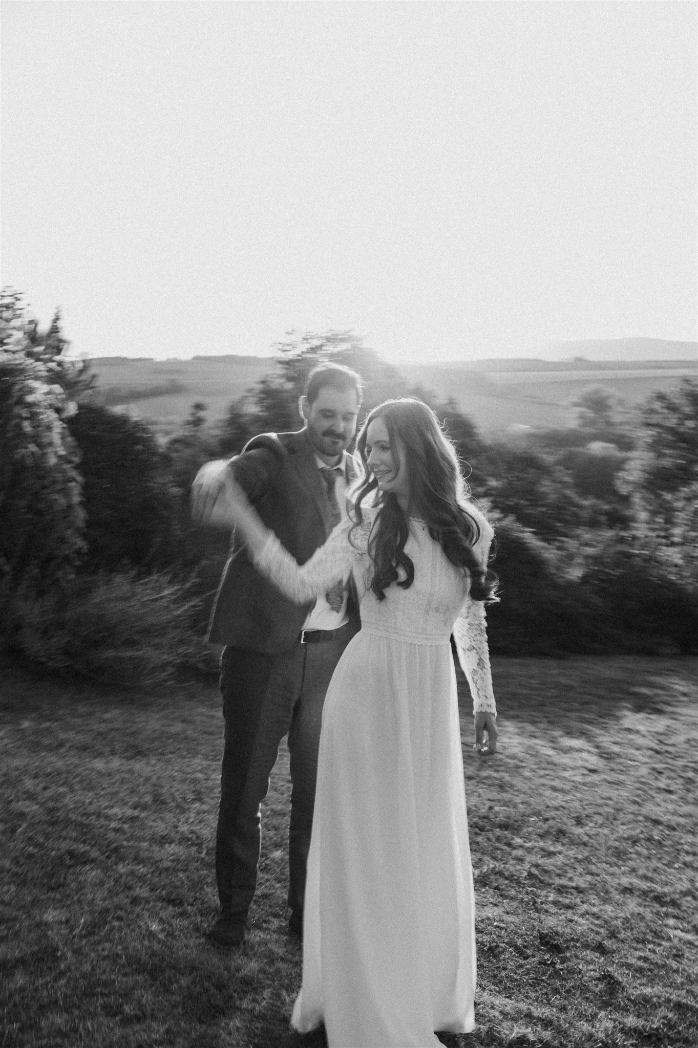 Black and white photo of a couple in formal attire dancing in a meadow at sunset.