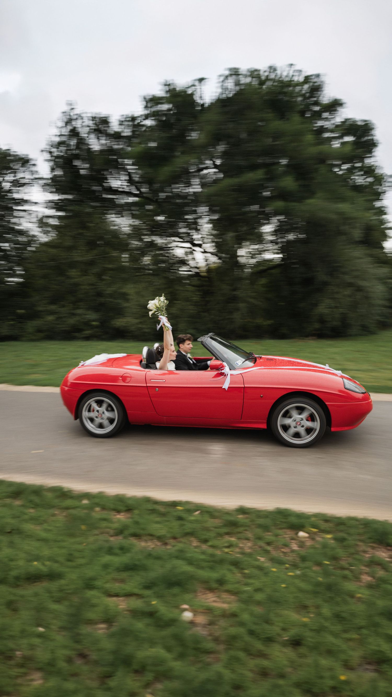 A sleek red convertible sports car drives along a winding road with trees in the background.