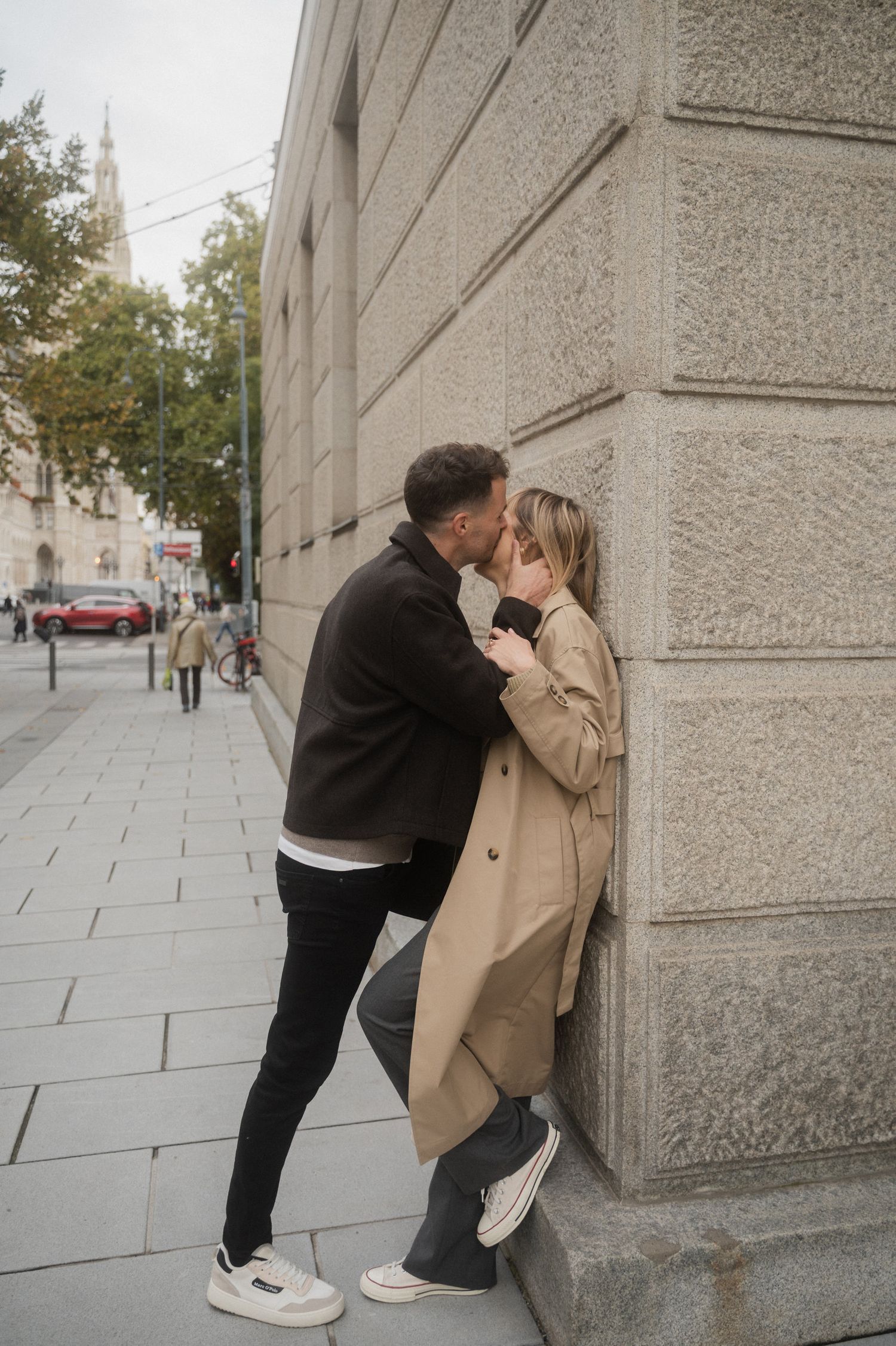 A romantic couple shares an intimate moment against a stone building wall on a city sidewalk.