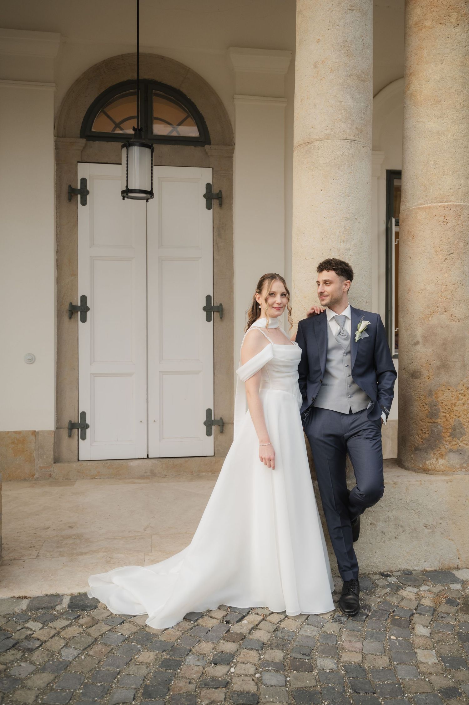 Bride in white gown and groom in navy suit posing by elegant stone columns at wedding venue.