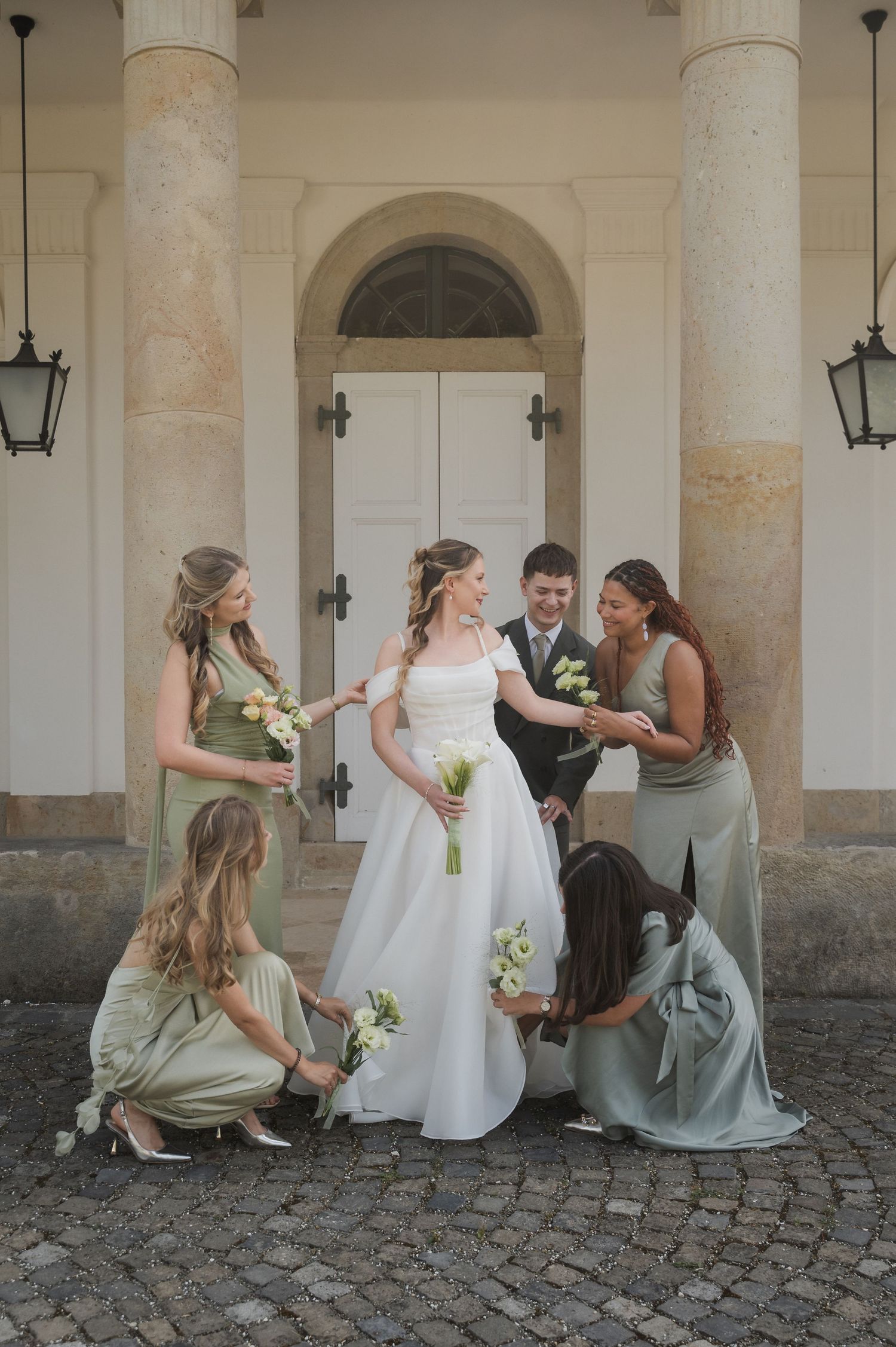 Bride in white gown surrounded by bridesmaids in sage green dresses outside elegant venue doors.