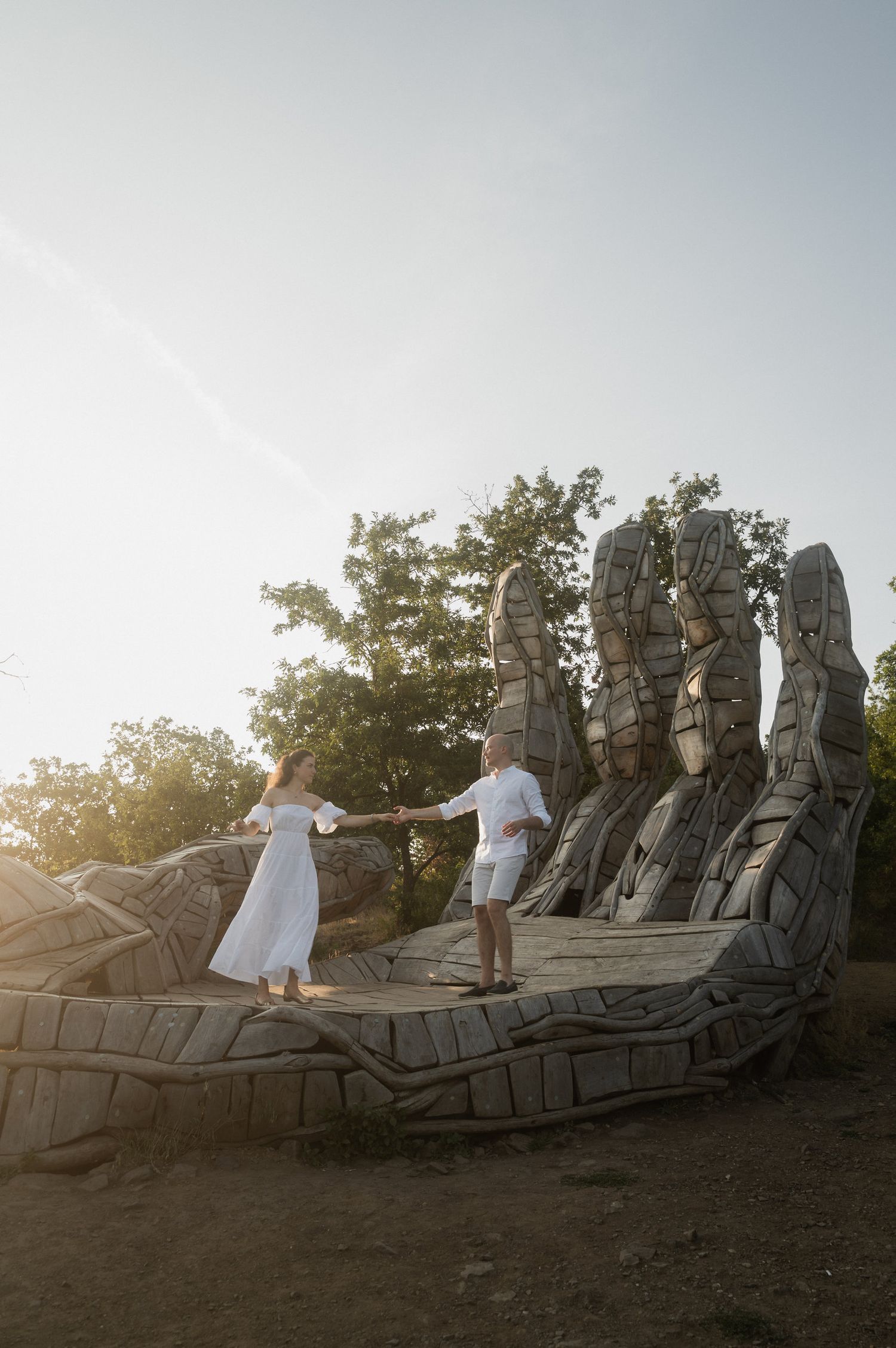 Two people in white outfits stand on a large wooden sculpture against a sunset sky with trees in the background.
