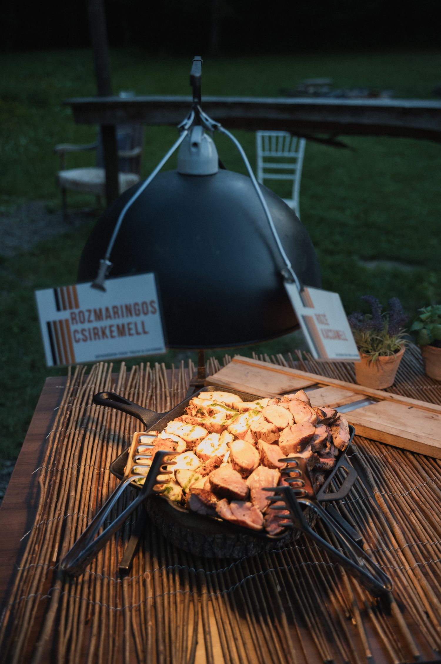 Grilled salmon fillets cooking on a wooden plank under a black grill dome in an outdoor setting at dusk.