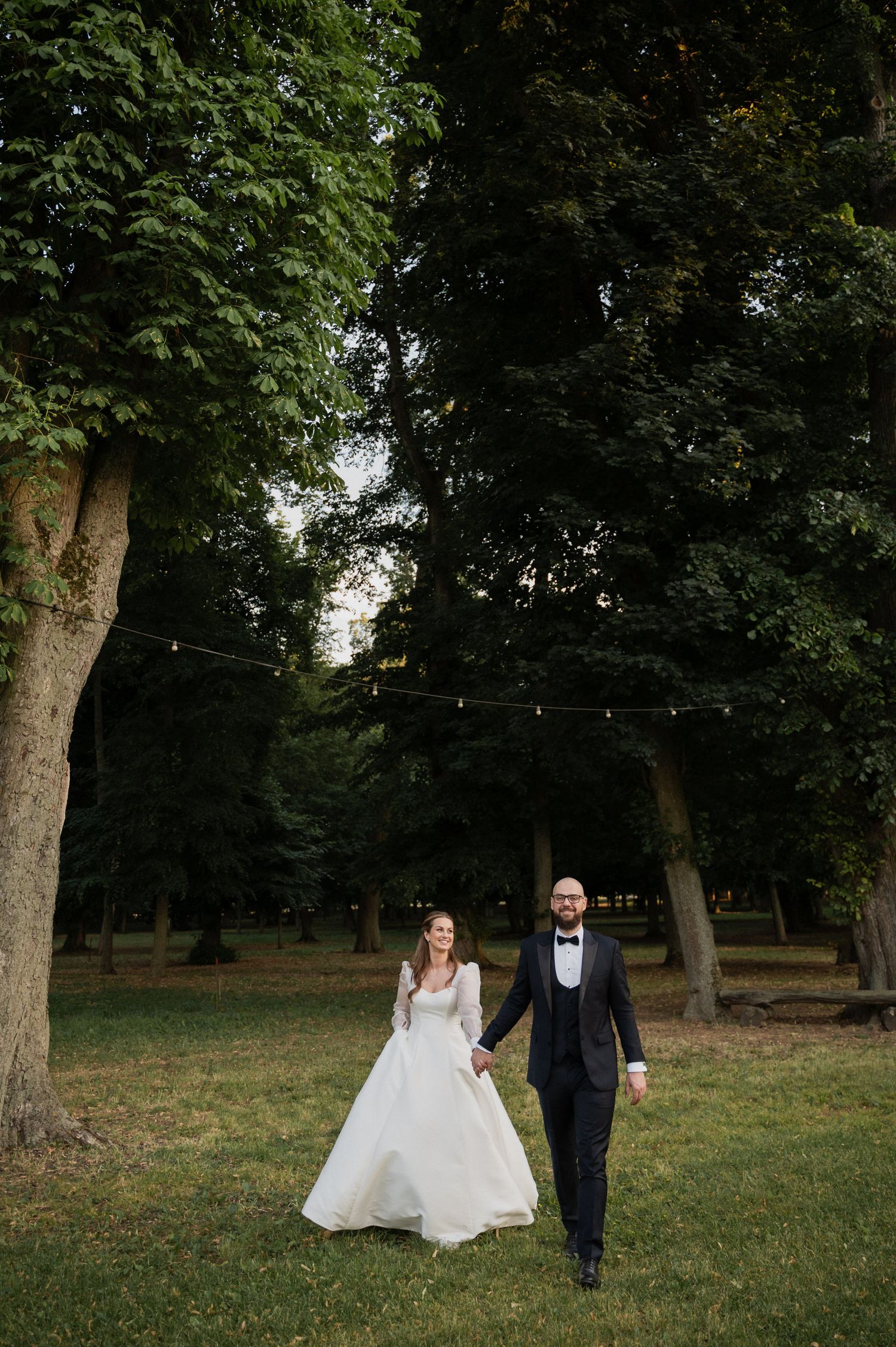 A couple in formal wedding attire walks together through a lush green park setting with tall trees in the background.