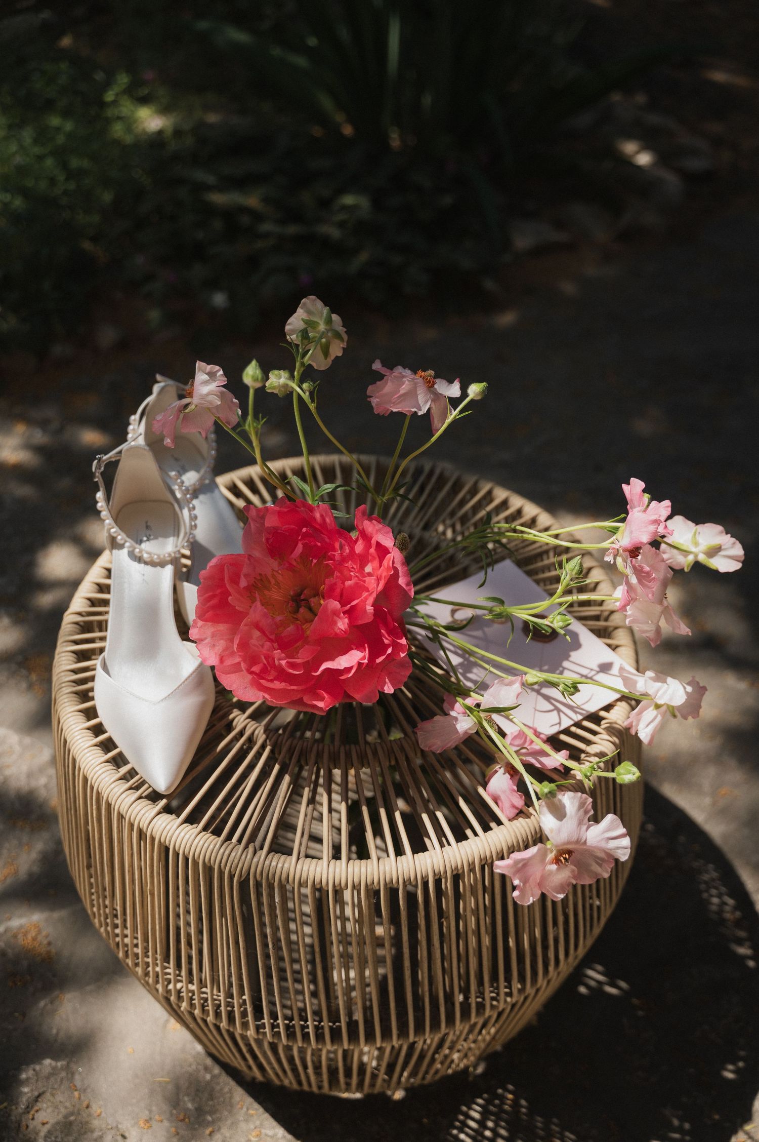 White bridal heels and pink flowers arranged in a wicker basket centerpiece outdoors.