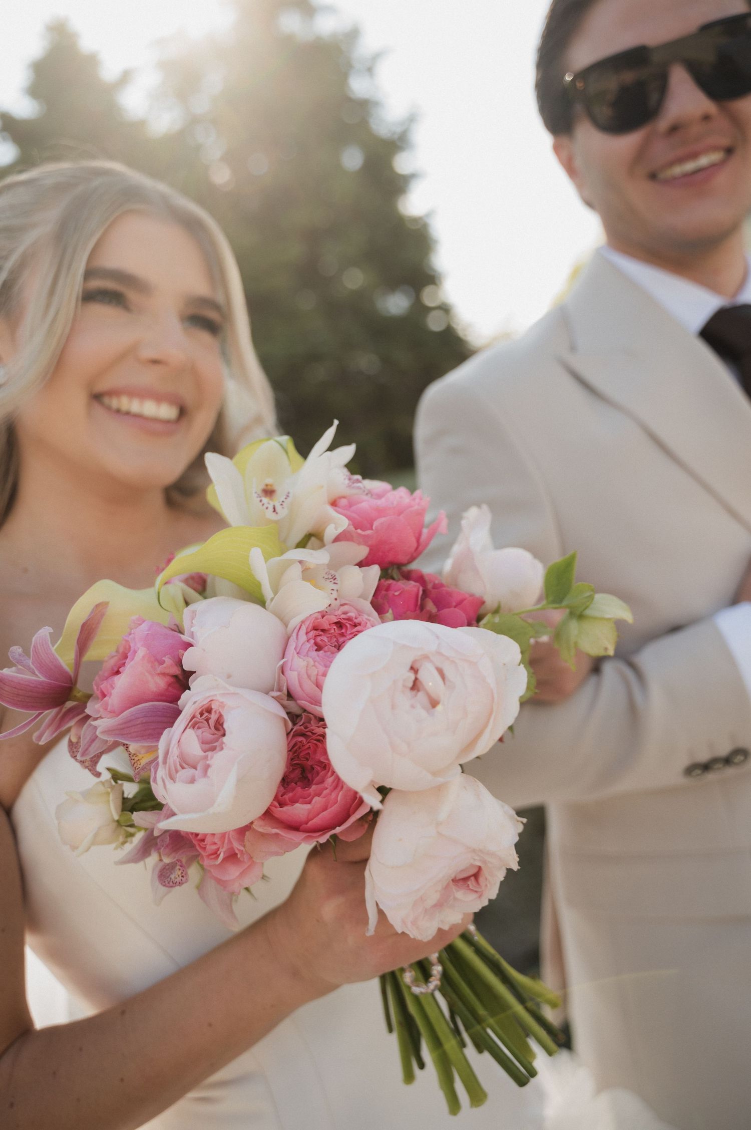Smiling bride holding a lush pink peony and rose bouquet beside groom in sunlit outdoor setting.