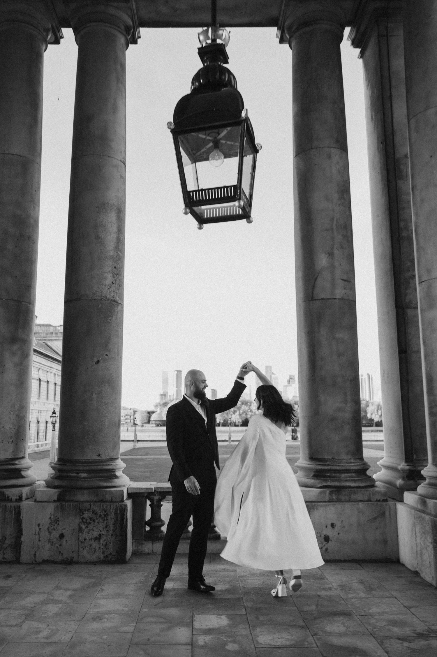 Wedding couple dances beneath historic colonnade with city view in background.